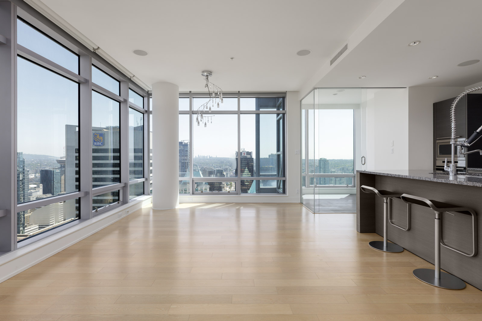 Modern high-rise apartment with large floor-to-ceiling windows, light wood floors, a kitchen area with bar stools, and city views.