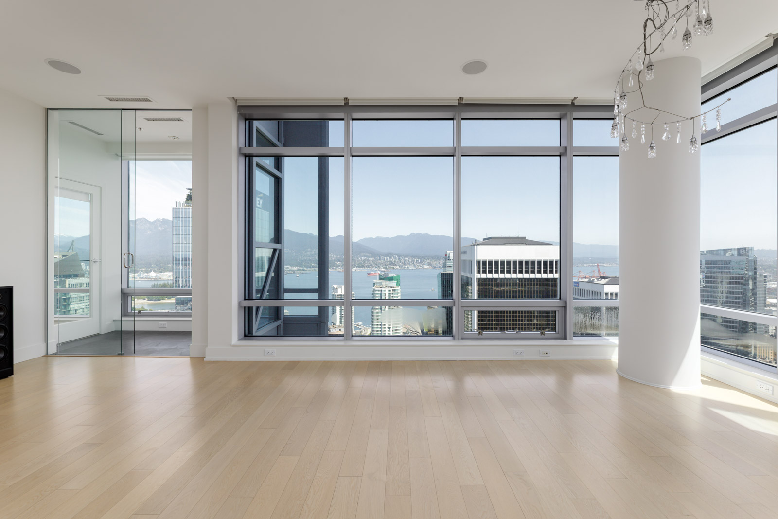 A modern, empty room with light wood floors, large floor-to-ceiling windows, city buildings, and mountains visible in the background.