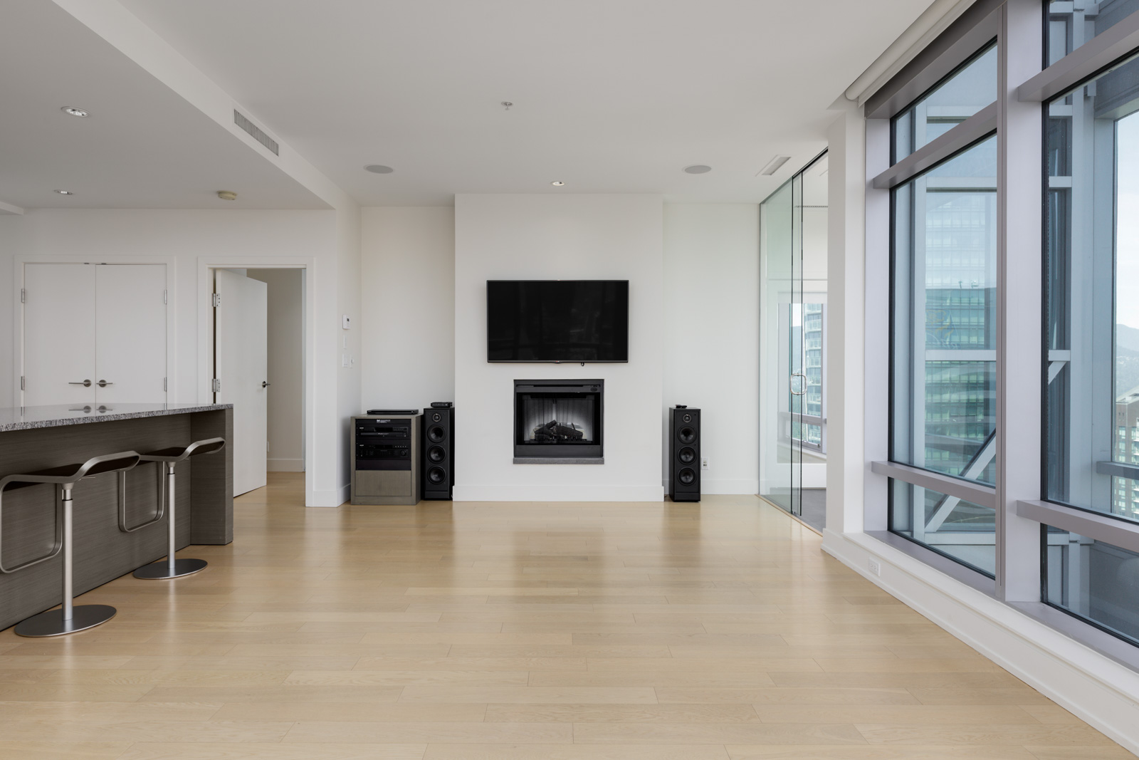 Modern living room with light wood floors, a wall-mounted TV above a fireplace, speakers, bar stools, and large floor-to-ceiling windows offering a city view.
