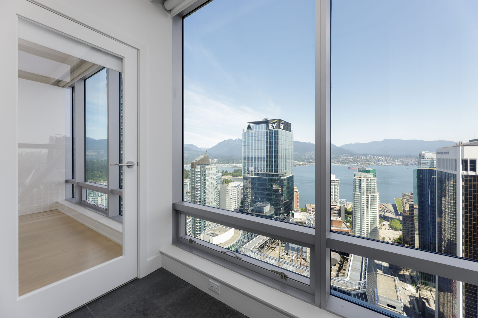 High-rise apartment room with large windows offering a view of city buildings, water, and distant mountains under a clear sky.