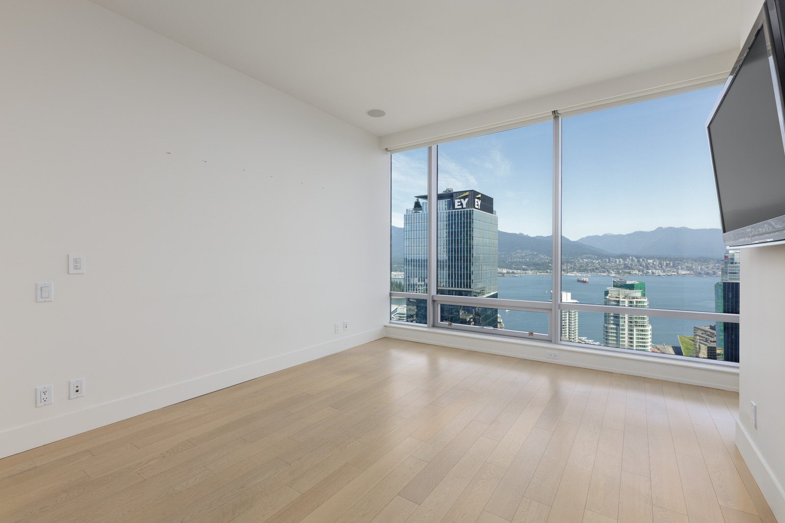 Empty room with light wood flooring, large windows showing city buildings and mountains, and a wall-mounted TV.