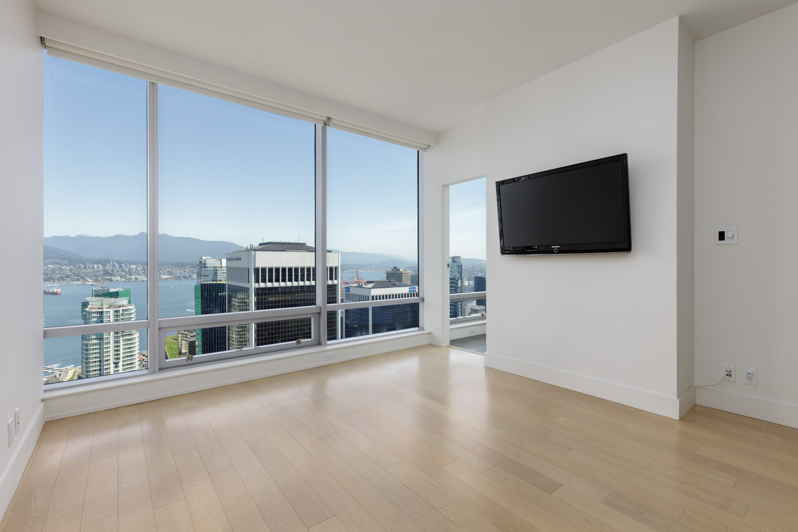 Modern empty room with light wood flooring, large floor-to-ceiling windows showcasing a city and mountain view, and a flat-screen TV mounted on a white wall.