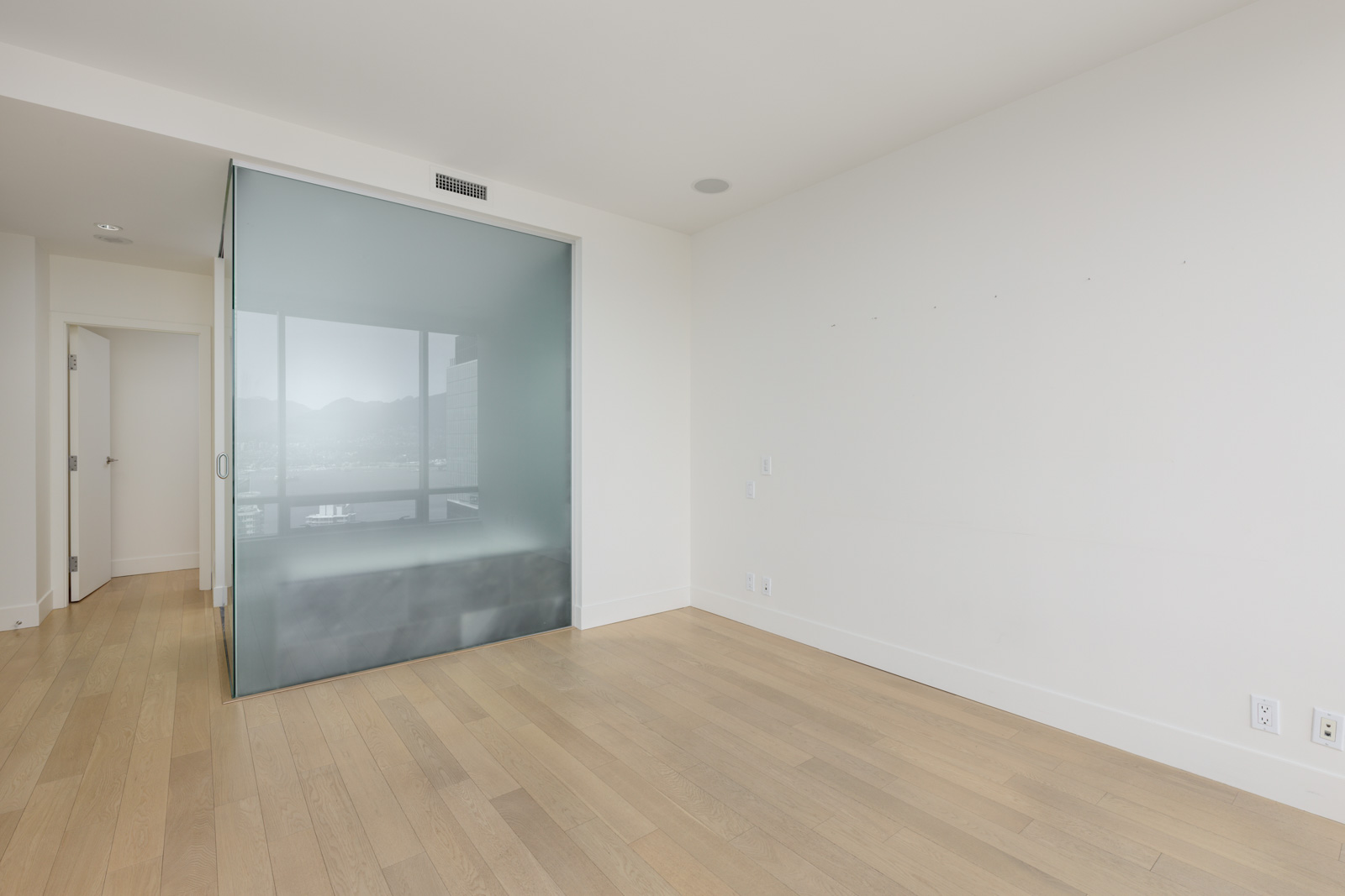Empty room with light wood flooring, white walls, and a frosted glass partition partially separating an adjacent area; doorway and natural light visible.