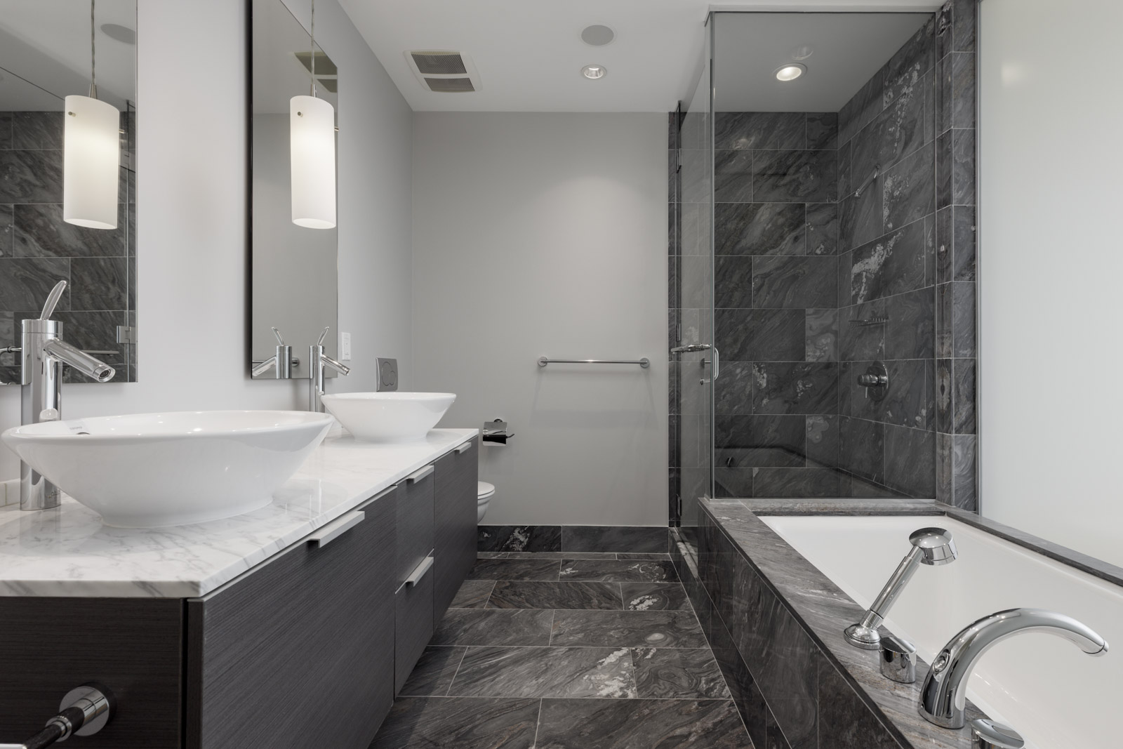 Modern bathroom with dark marble tiles, double sinks on a marble countertop, glass-enclosed shower, and a bathtub with chrome fixtures.