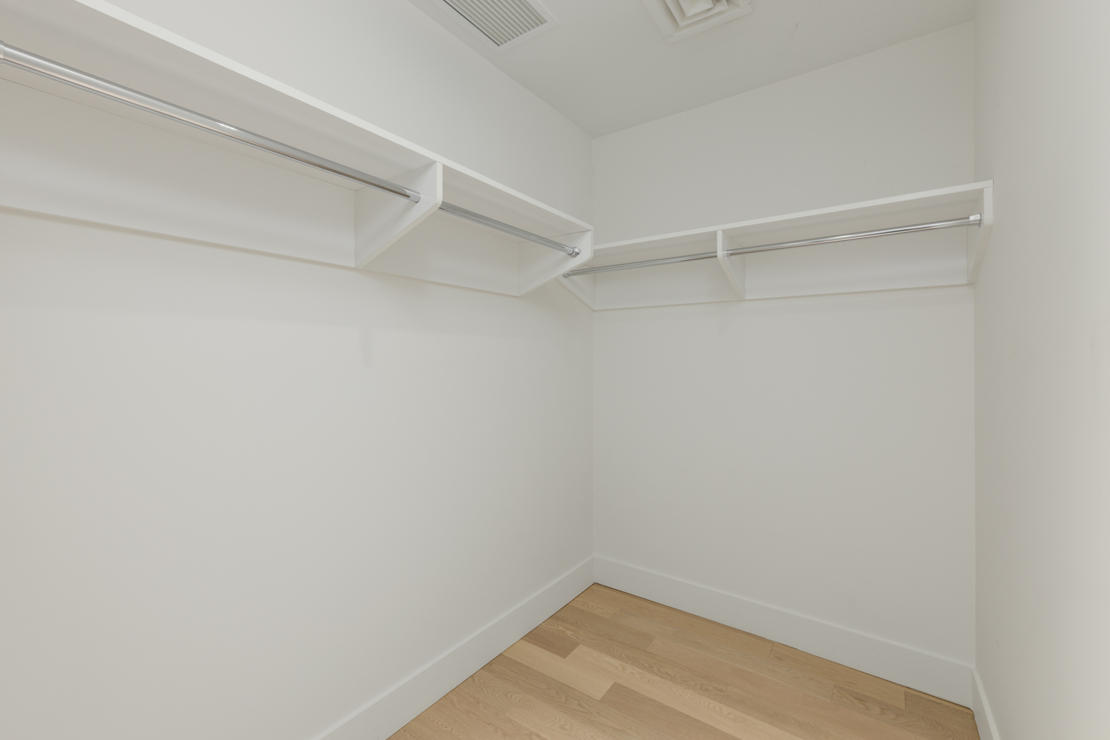 Empty walk-in closet with light wood flooring, white walls, and metal hanging rods with shelves along two walls.