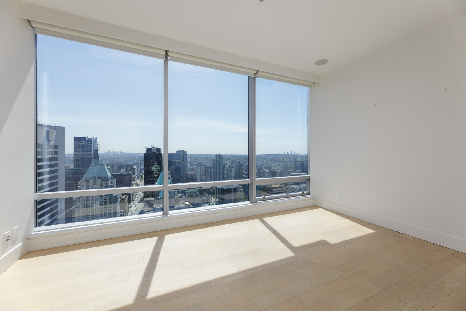 Empty room with large floor-to-ceiling windows showing a city skyline view, white walls, and light wood flooring.