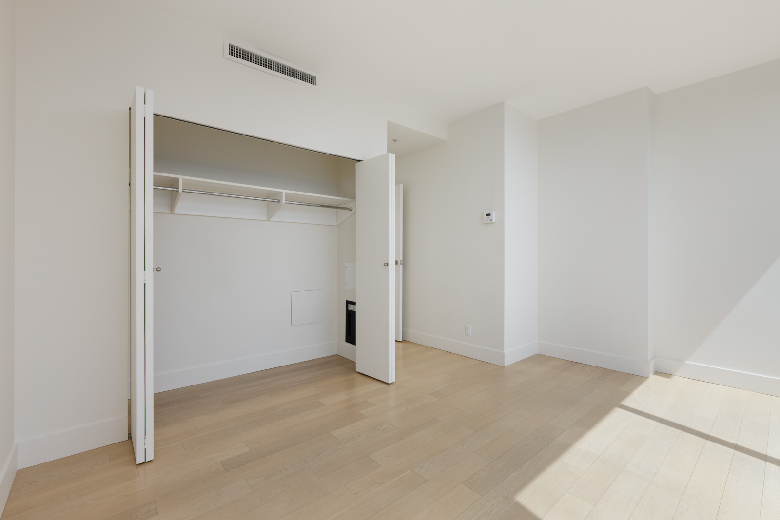 Empty room with light wood flooring, white walls, and an open closet with shelves and hanging rod. Sunlight streams in through a window out of frame.