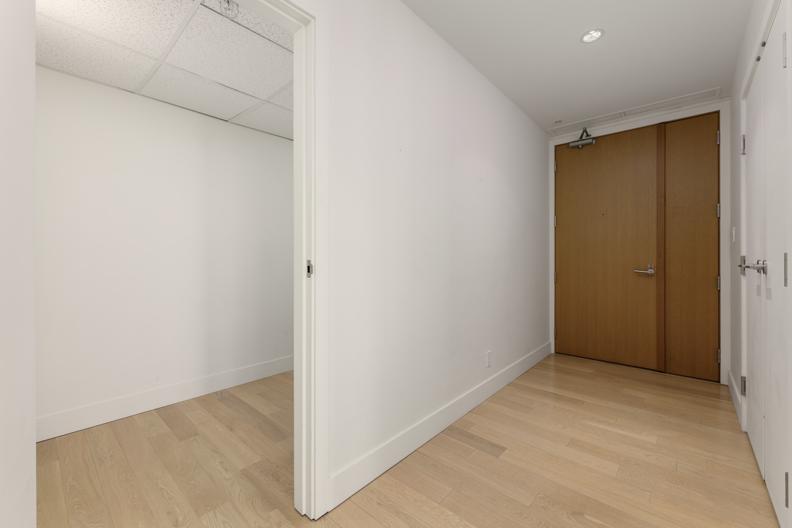 A hallway with light wood flooring, white walls, a wooden entrance door, and an adjacent small room with a drop ceiling.