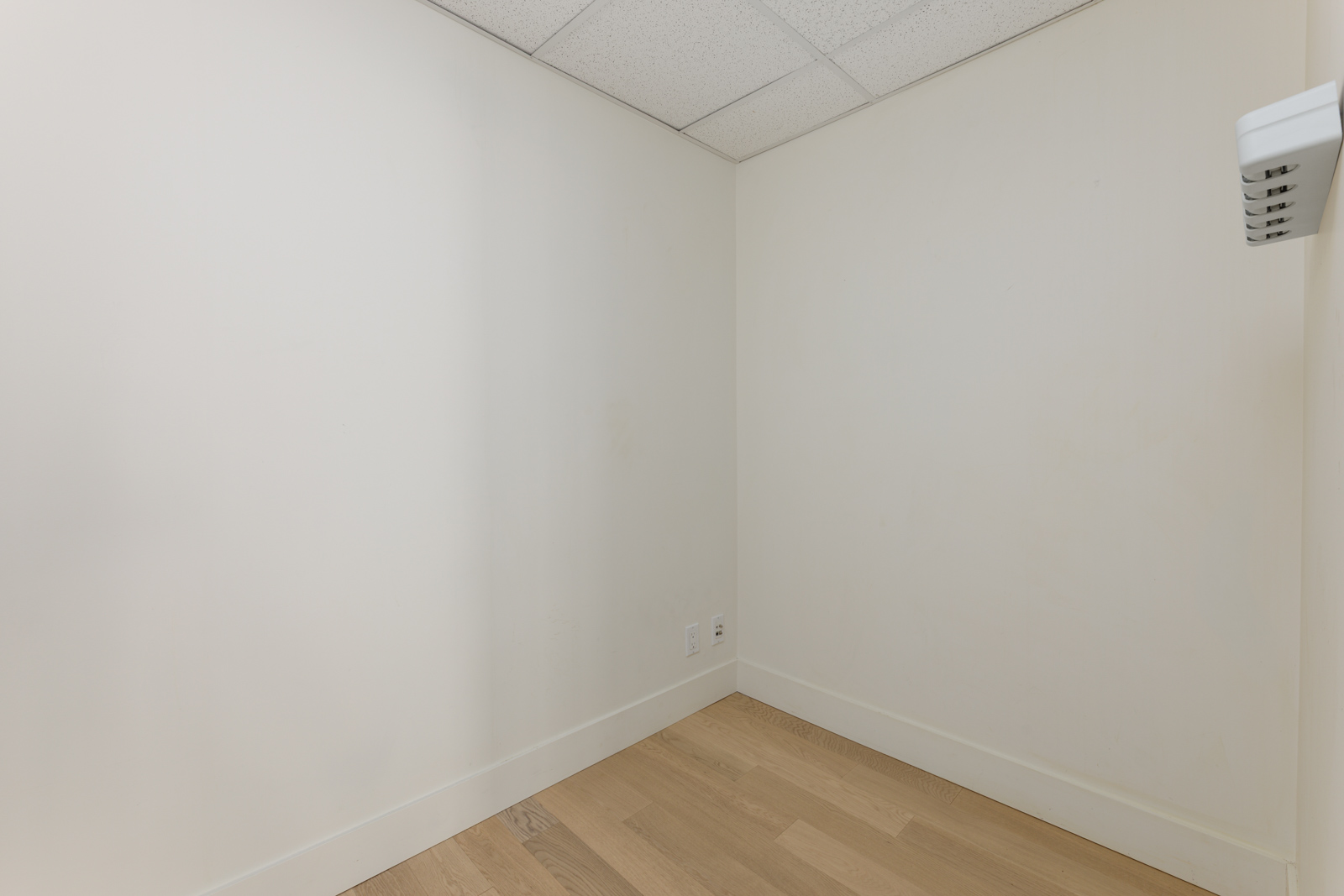 Small, empty room with off-white walls, light wood flooring, a white ceiling with tiles, and a wall outlet near the floor.