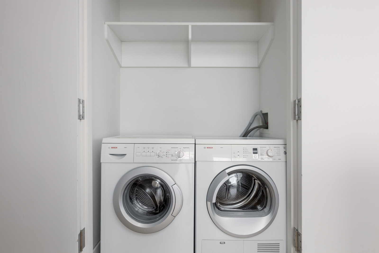A white front-load washing machine and dryer are placed side by side in a small laundry closet with open shelves above them.