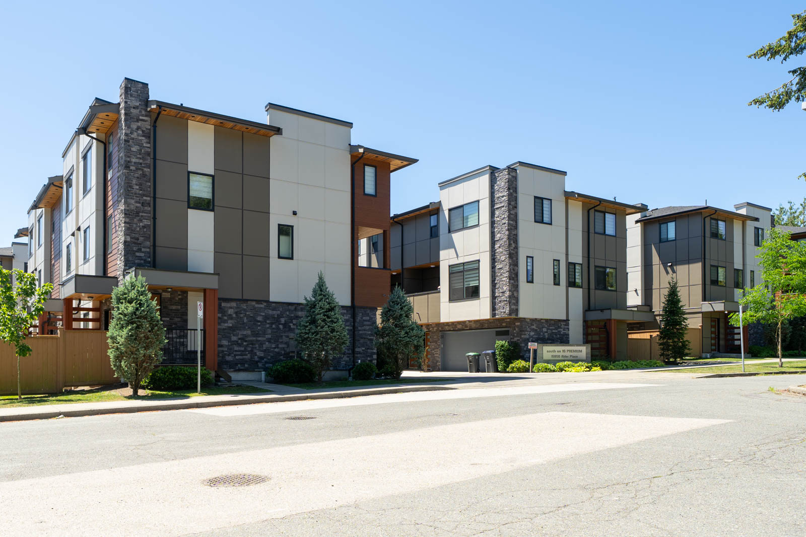 Modern three-story townhouse buildings with flat roofs and large windows, set along a quiet street with trees and shrubs in front.