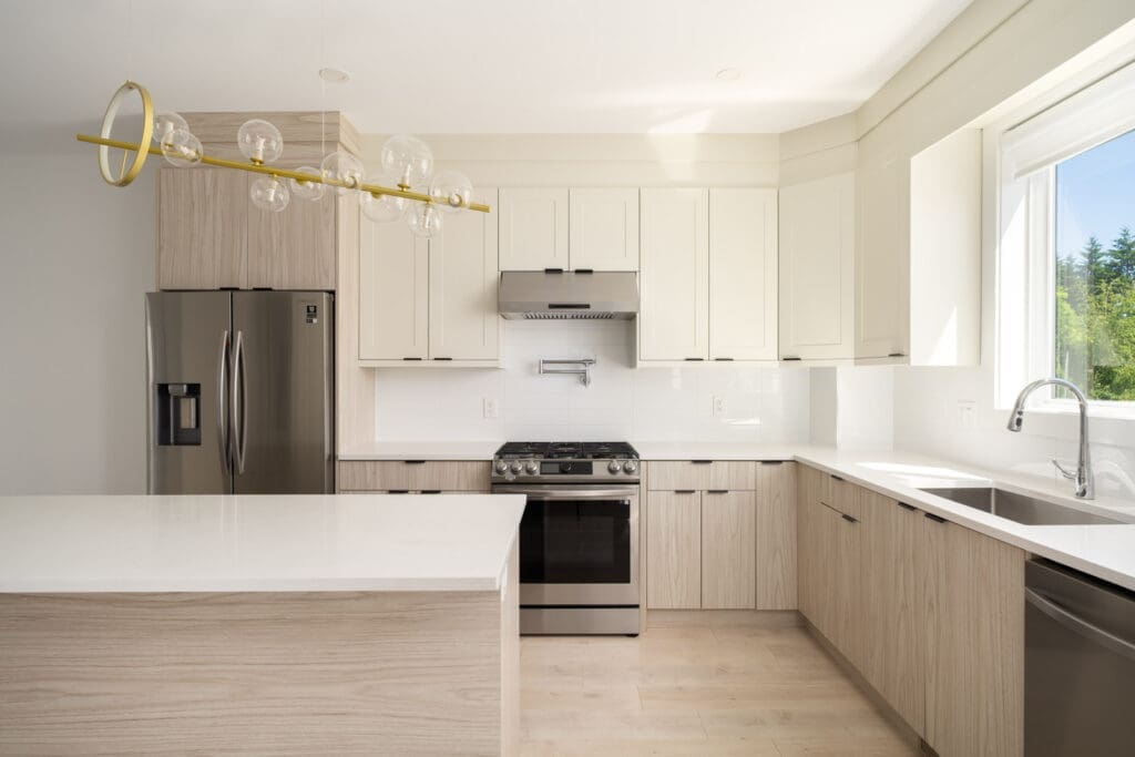 Modern kitchen with light wood cabinets, white countertops, stainless steel appliances, large window, and a contemporary light fixture.