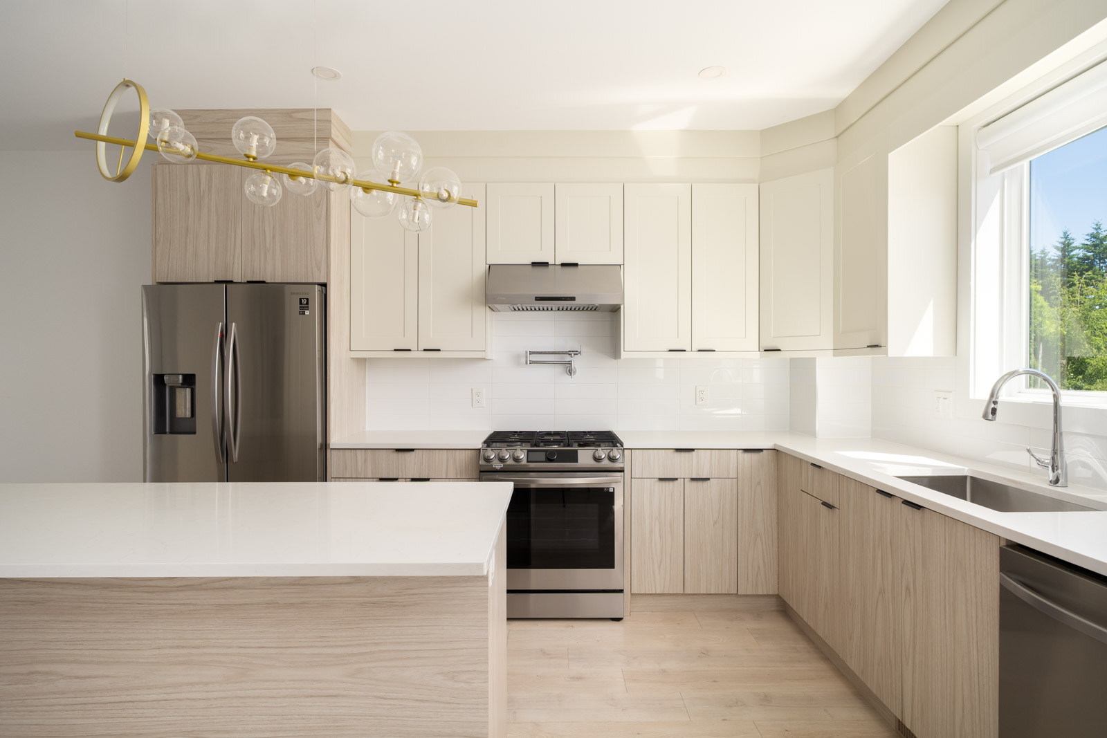 Modern kitchen with light wood cabinets, white countertops, stainless steel appliances, large window, and a contemporary light fixture.