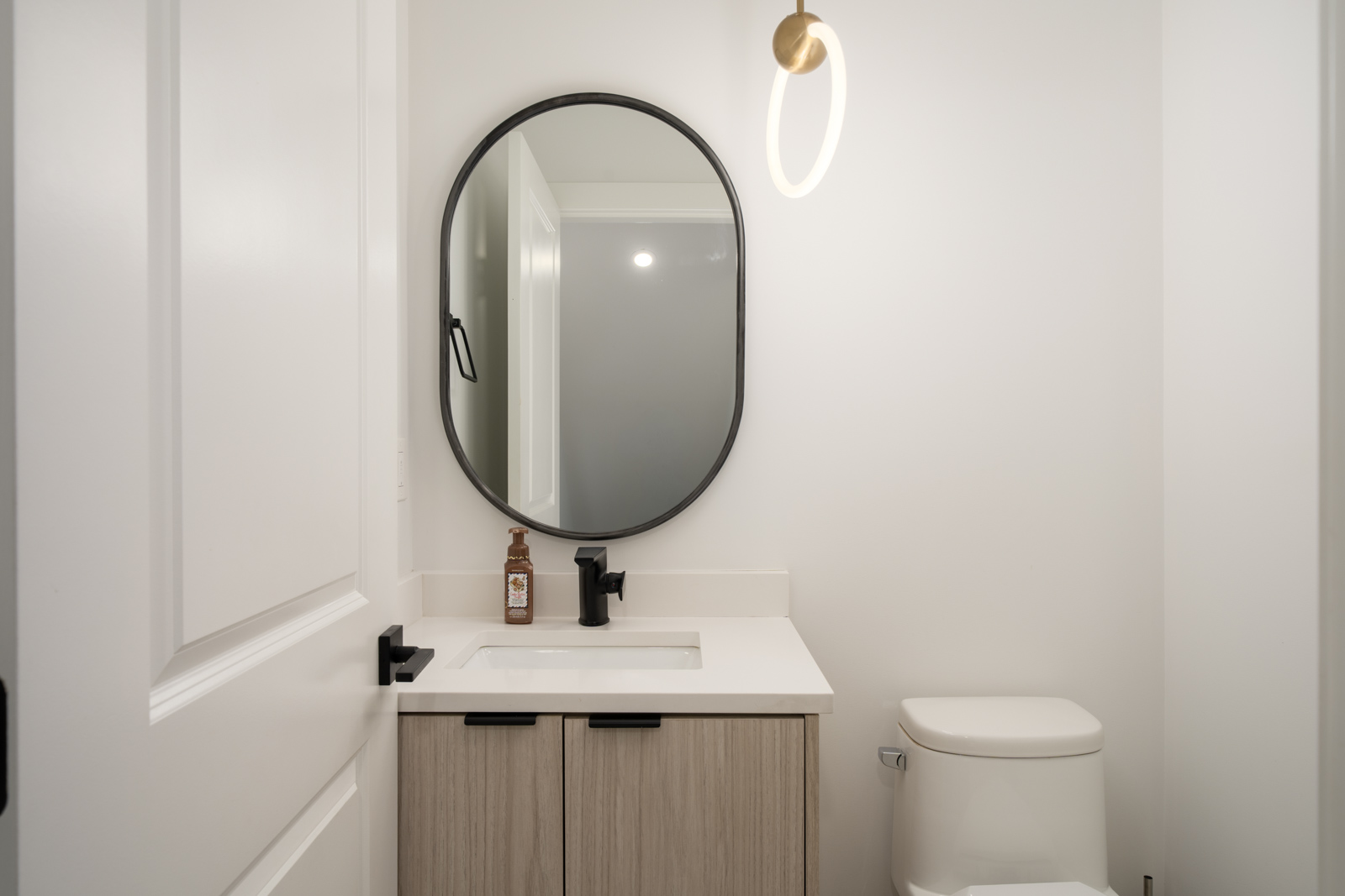 Modern bathroom with a round black-framed mirror, single sink vanity with light wood cabinets, a black faucet, a wall sconce, and a white toilet against white walls.