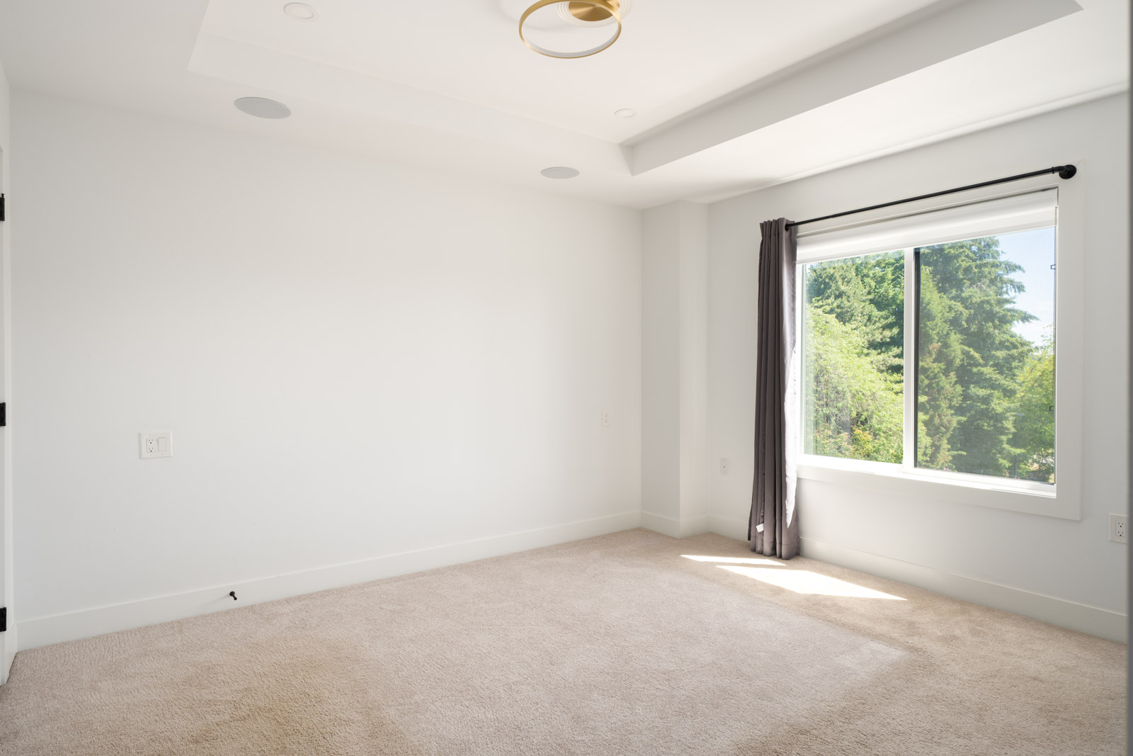 Empty bedroom with beige carpet, white walls, a window with gray curtains, and a ceiling light fixture. Sunlight streams in through the window, illuminating the room.