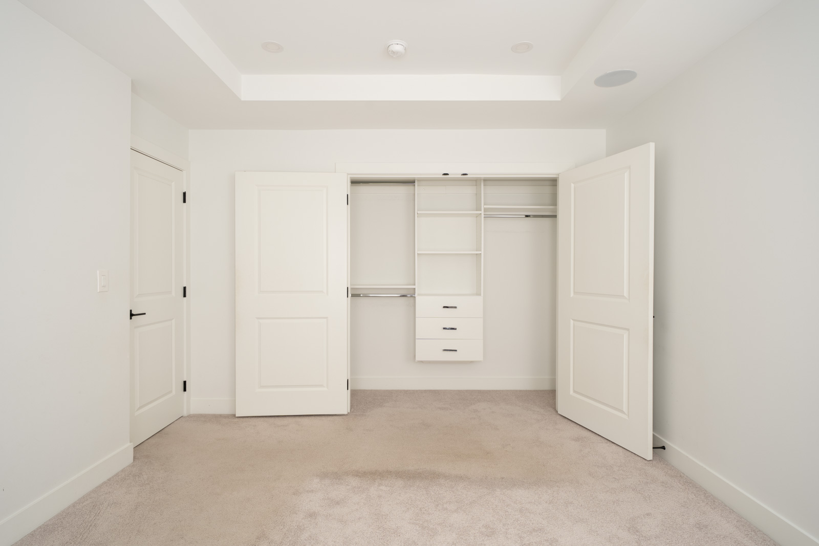Empty room with beige carpet, white walls, and open double closet doors revealing shelves, drawers, and hanging rods inside.