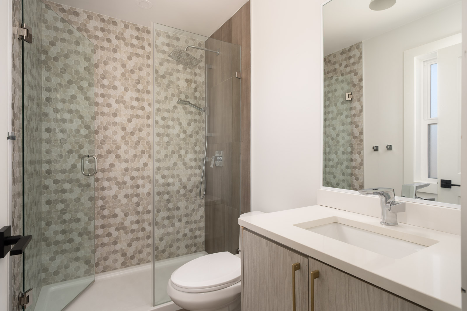 Modern bathroom with a glass-enclosed shower featuring hexagonal tiles, a white countertop with integrated sink, and light wood cabinets.