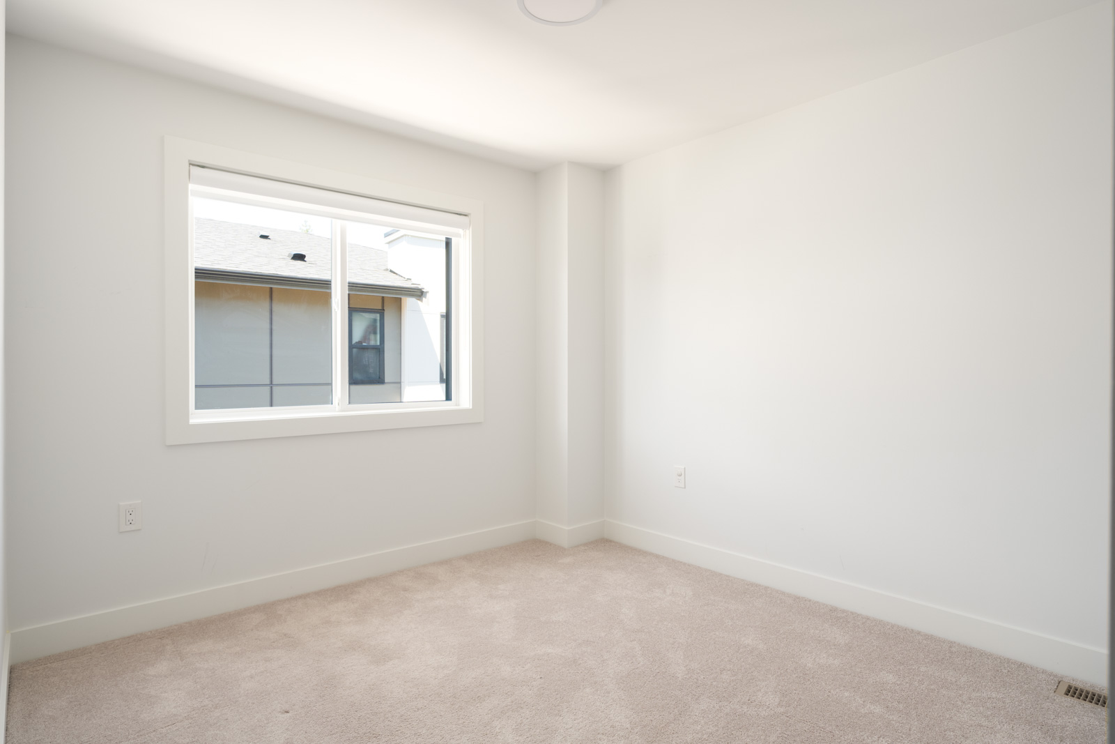 Empty room with white walls, beige carpet, a single window letting in daylight, and minimal corners and power outlets visible.