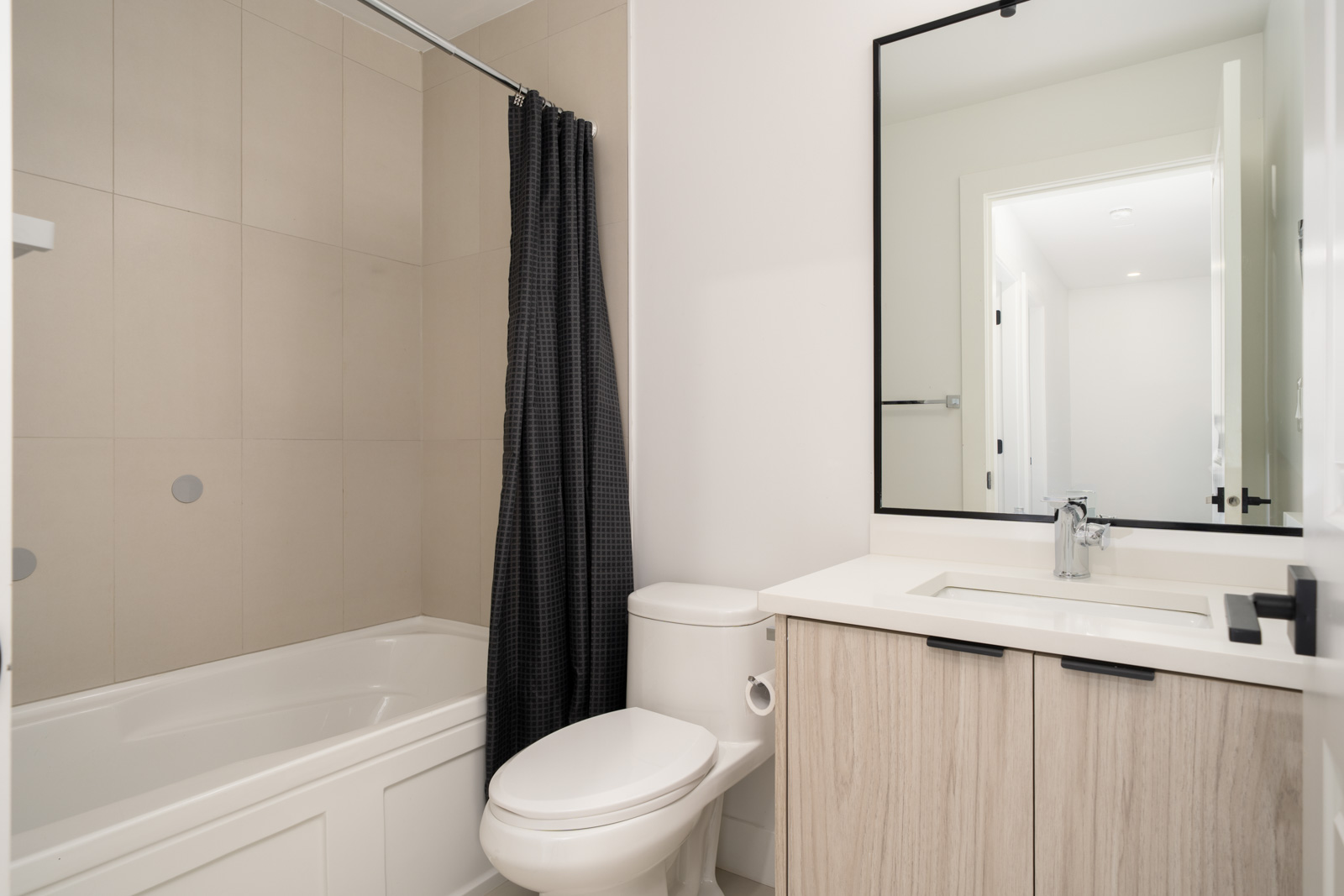 Modern bathroom with a bathtub-shower combo, toilet, light wood vanity, rectangular mirror, and black shower curtain. Walls are white and floor is light-colored tile.
