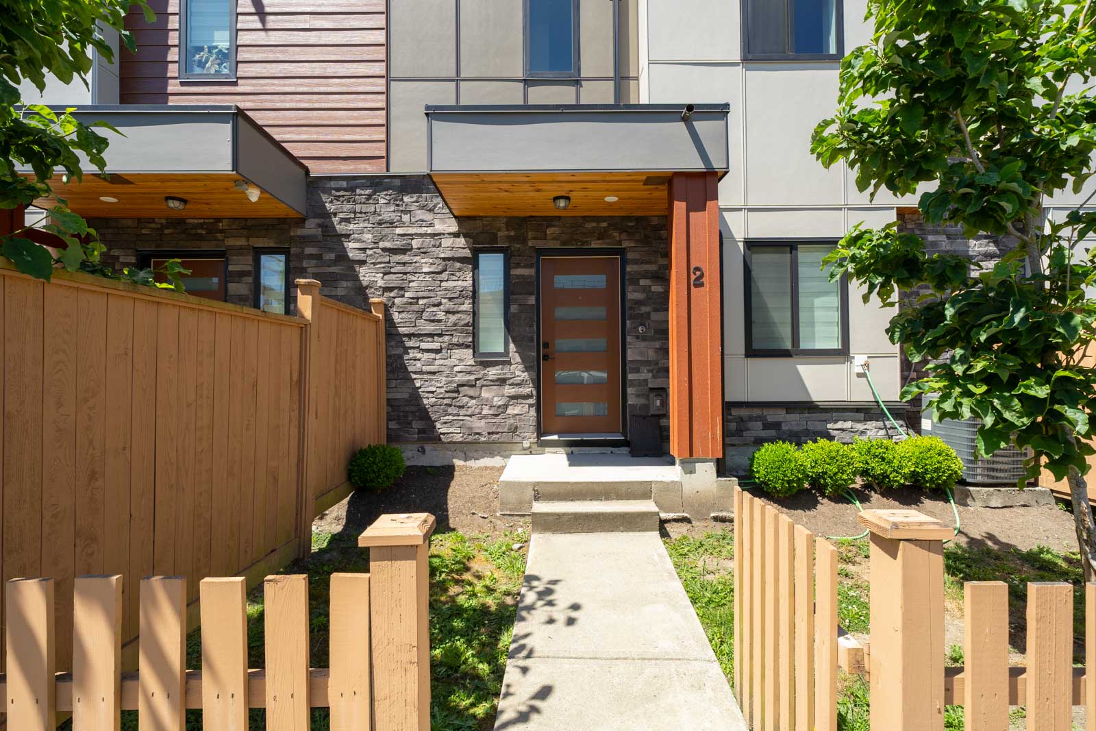 A modern townhouse with a wooden fence, stone and wood facade, and a glass-panel front door, viewed from the front yard path on a sunny day.