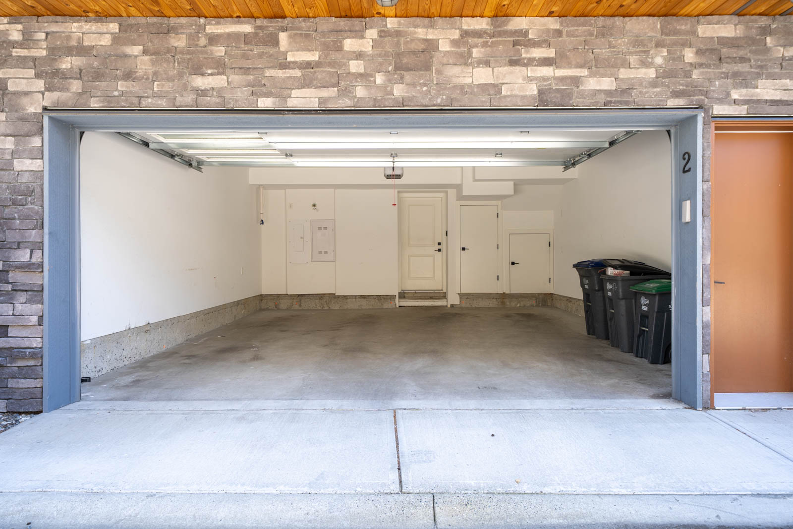 Open two-car garage with a clean concrete floor, white walls, utility panels, storage areas, and two trash bins along the right wall.