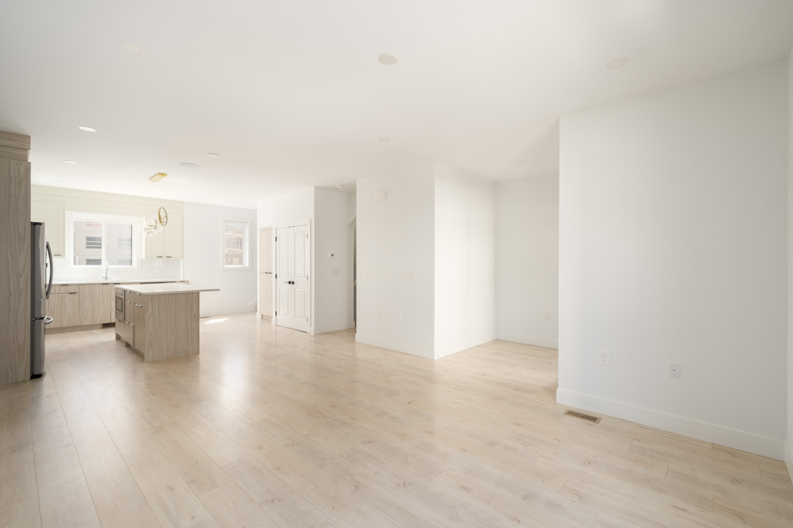 Unfurnished, modern open-plan living space with light wood floors, white walls, and a kitchen featuring stainless steel appliances and an island.