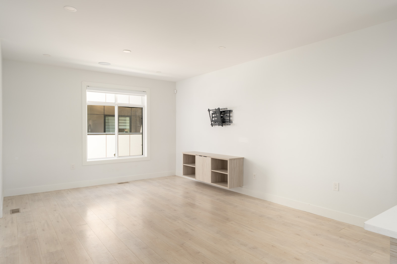 Empty living room with light wood flooring, a wall-mounted TV bracket, a small wooden media console, and a large window letting in natural light.