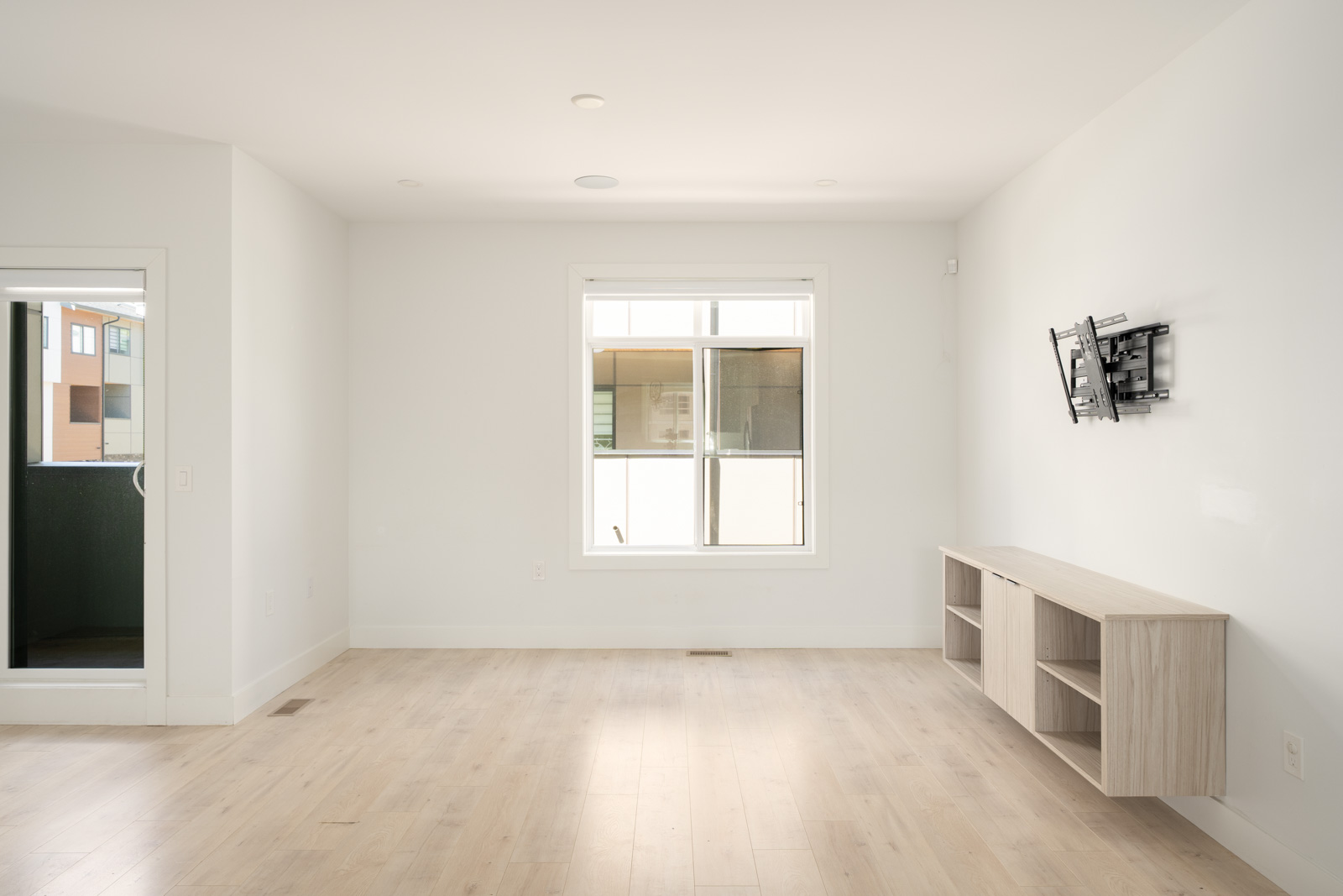 A bright, empty room with light wood flooring, a large window, a door to a balcony, a wall-mounted TV bracket, and a wooden shelf unit below.