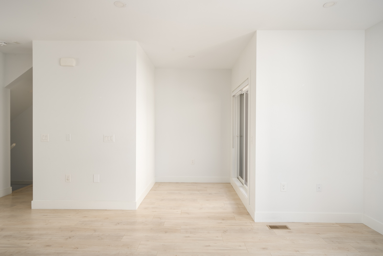 Empty room with white walls, light wood flooring, and a sliding glass door on the right side. The space is well-lit with natural light.