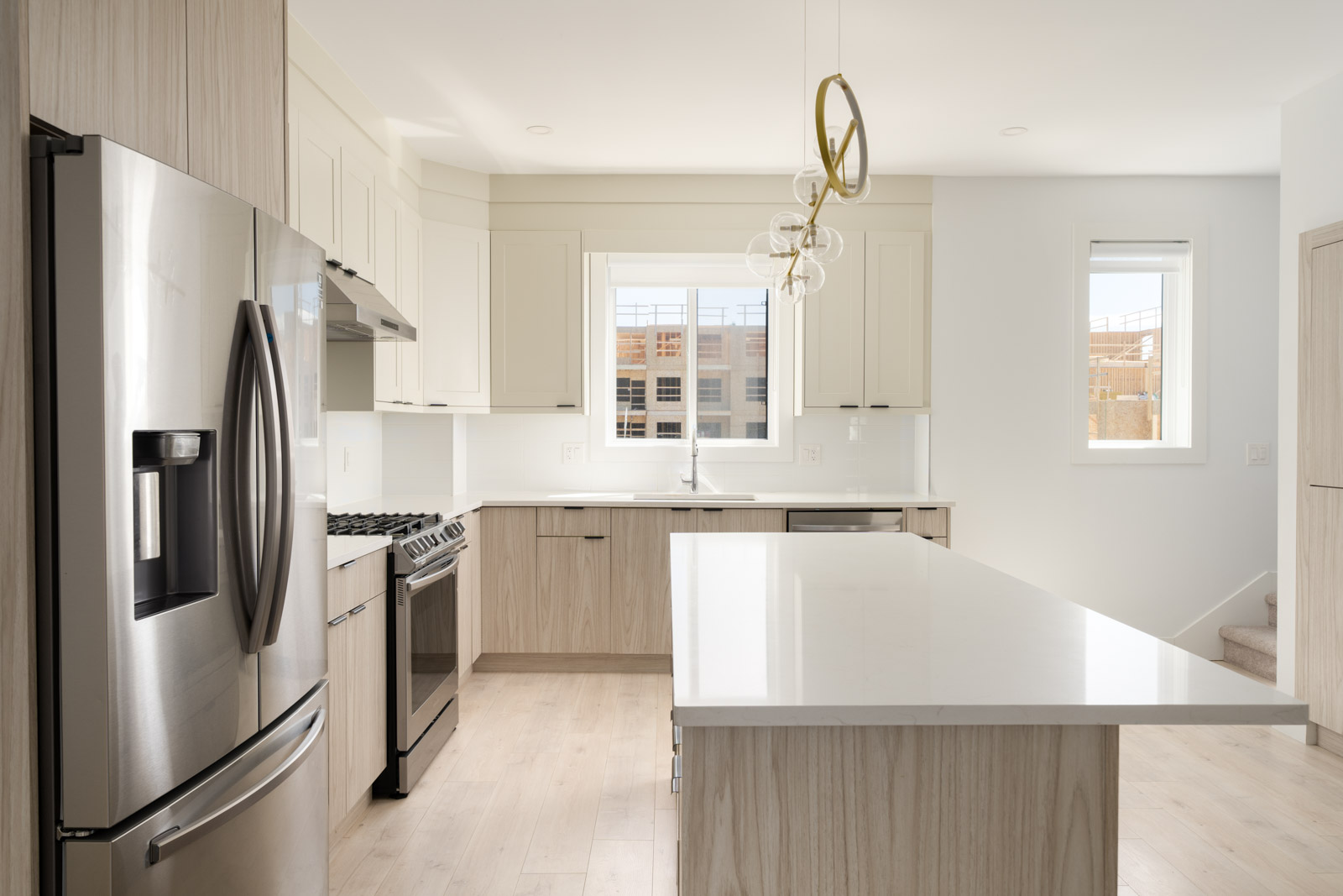 Modern kitchen with light wood cabinets, stainless steel appliances, a large central island, white countertops, and two windows letting in natural light.