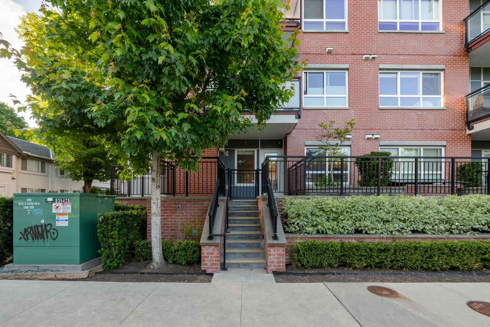 A brick apartment building with large windows, a central entrance up a short stairway, landscaped shrubs, and a green utility box on the left side of the sidewalk.