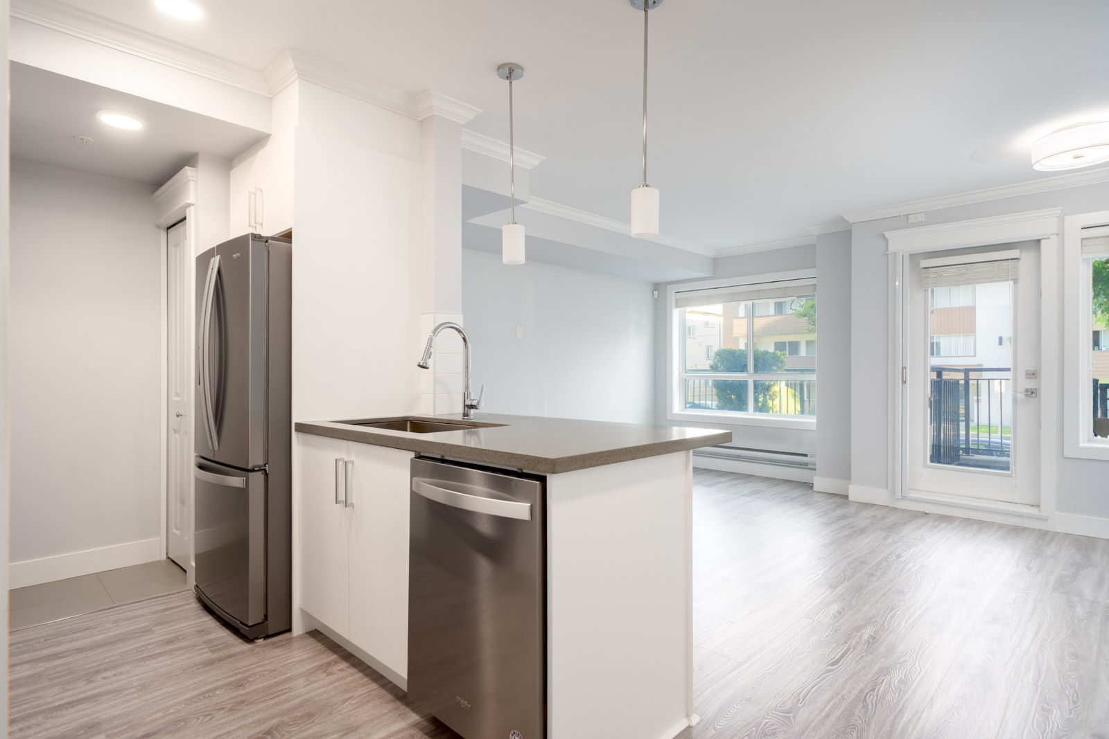 Modern kitchen with stainless steel appliances, white cabinetry, an island with sink, and open living area with large windows and light wood flooring.
