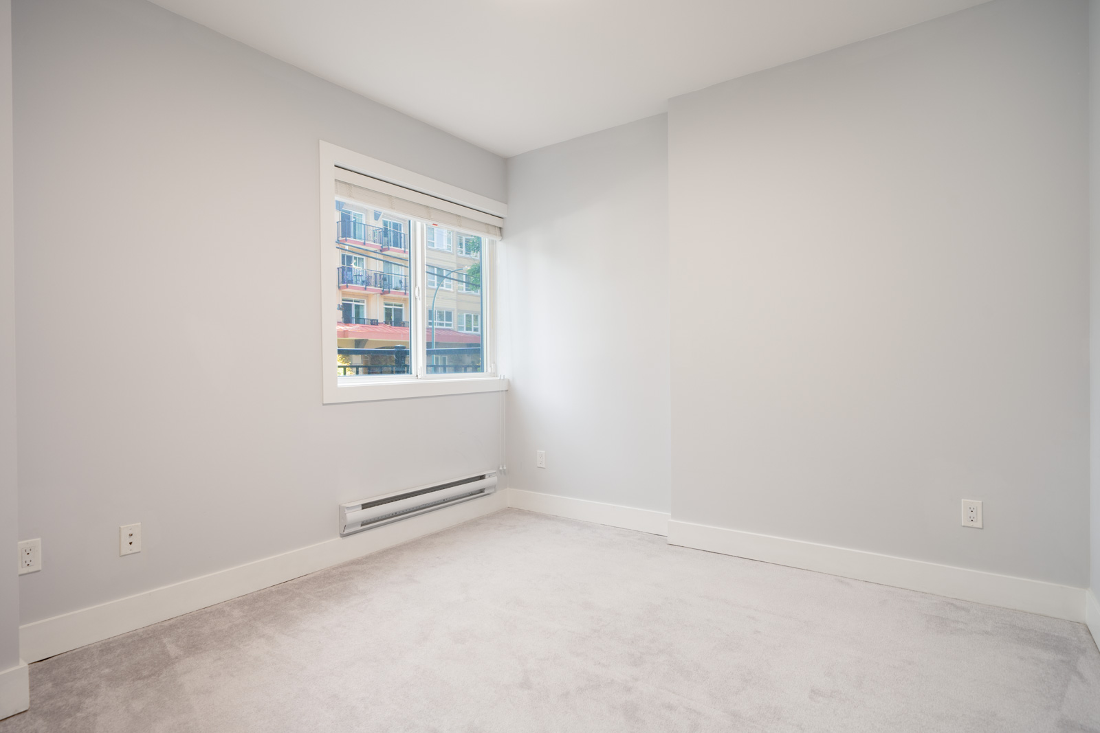 Empty room with light gray walls, white trim, carpeted floor, a window with a view of colorful buildings, and several electrical outlets.