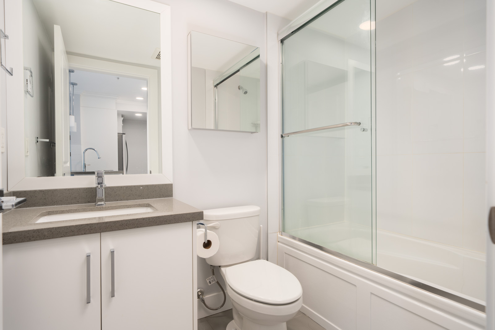 Modern bathroom with a white toilet, sink vanity with gray countertop, mounted mirror, and a glass-enclosed bathtub with shower.