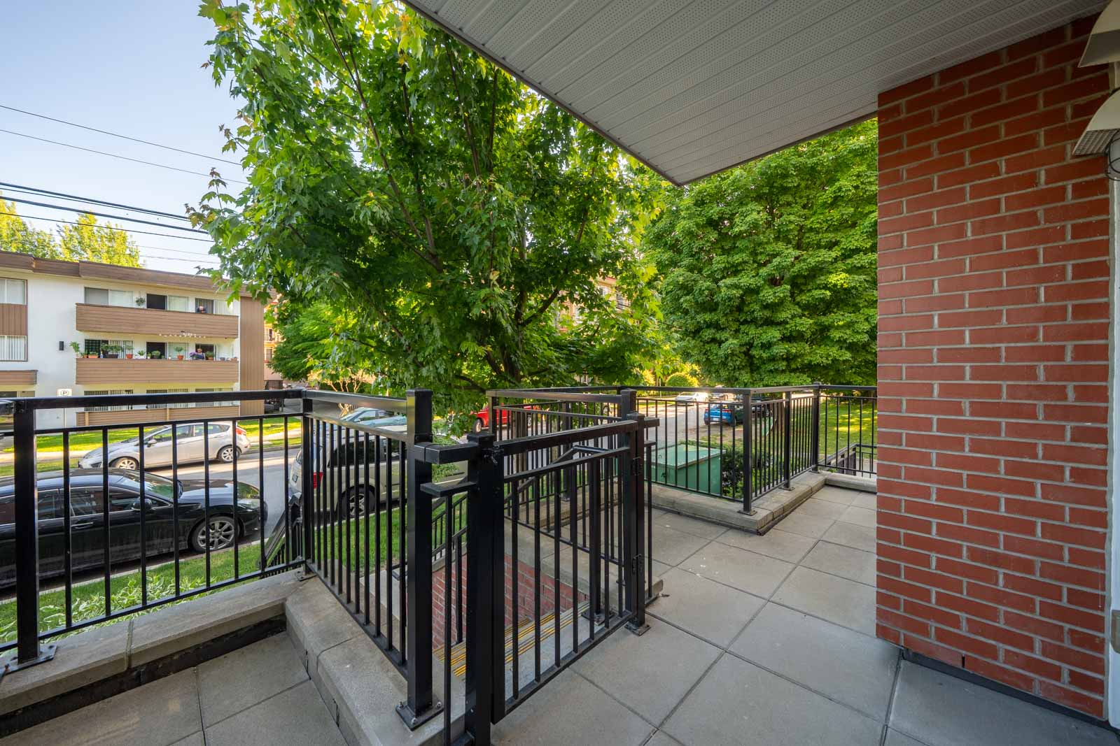 A ground-level patio with metal railings, brick wall, and tiled flooring overlooks a street with parked cars and trees in the background.