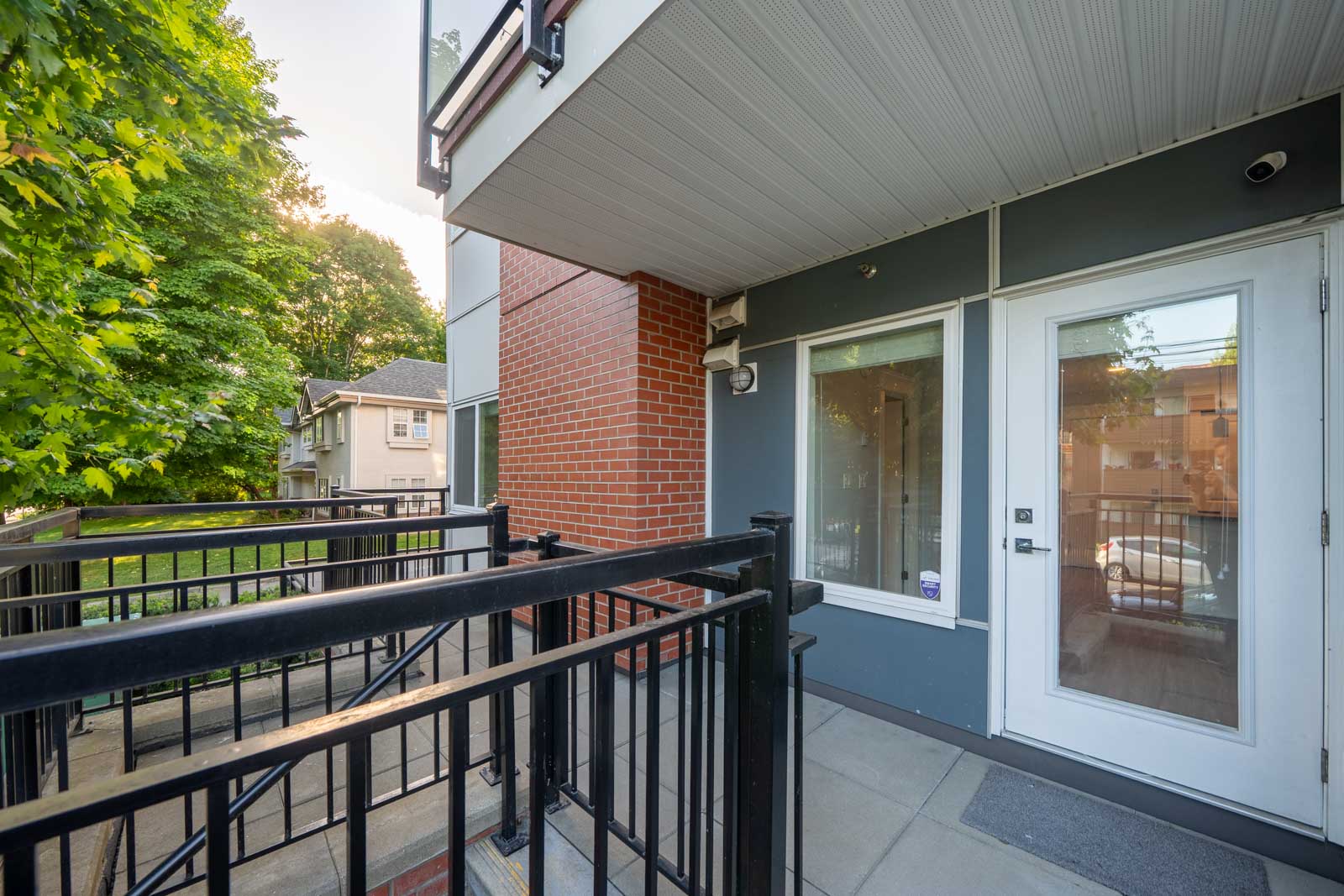 Ground-level apartment entry with a white door, glass panel, black metal railings, red brick column, and trees visible in the background.