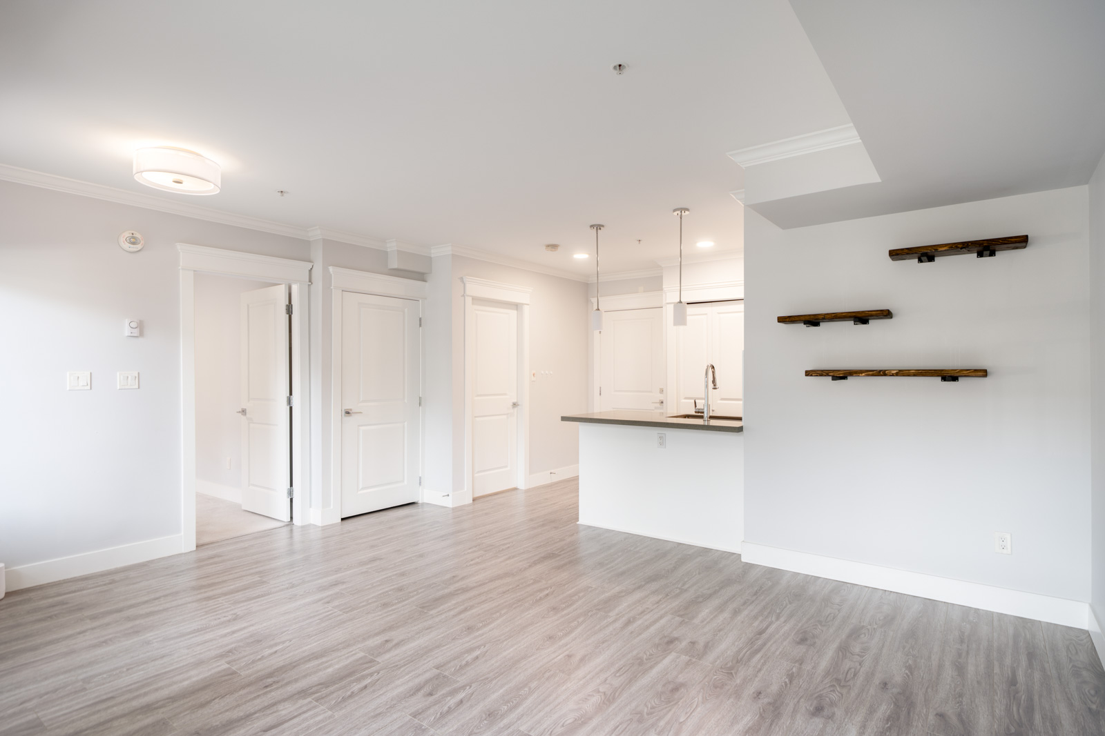 A modern, unfurnished apartment with light gray walls, wood-look flooring, a kitchen with a breakfast bar, and dark wood floating shelves on the wall.
