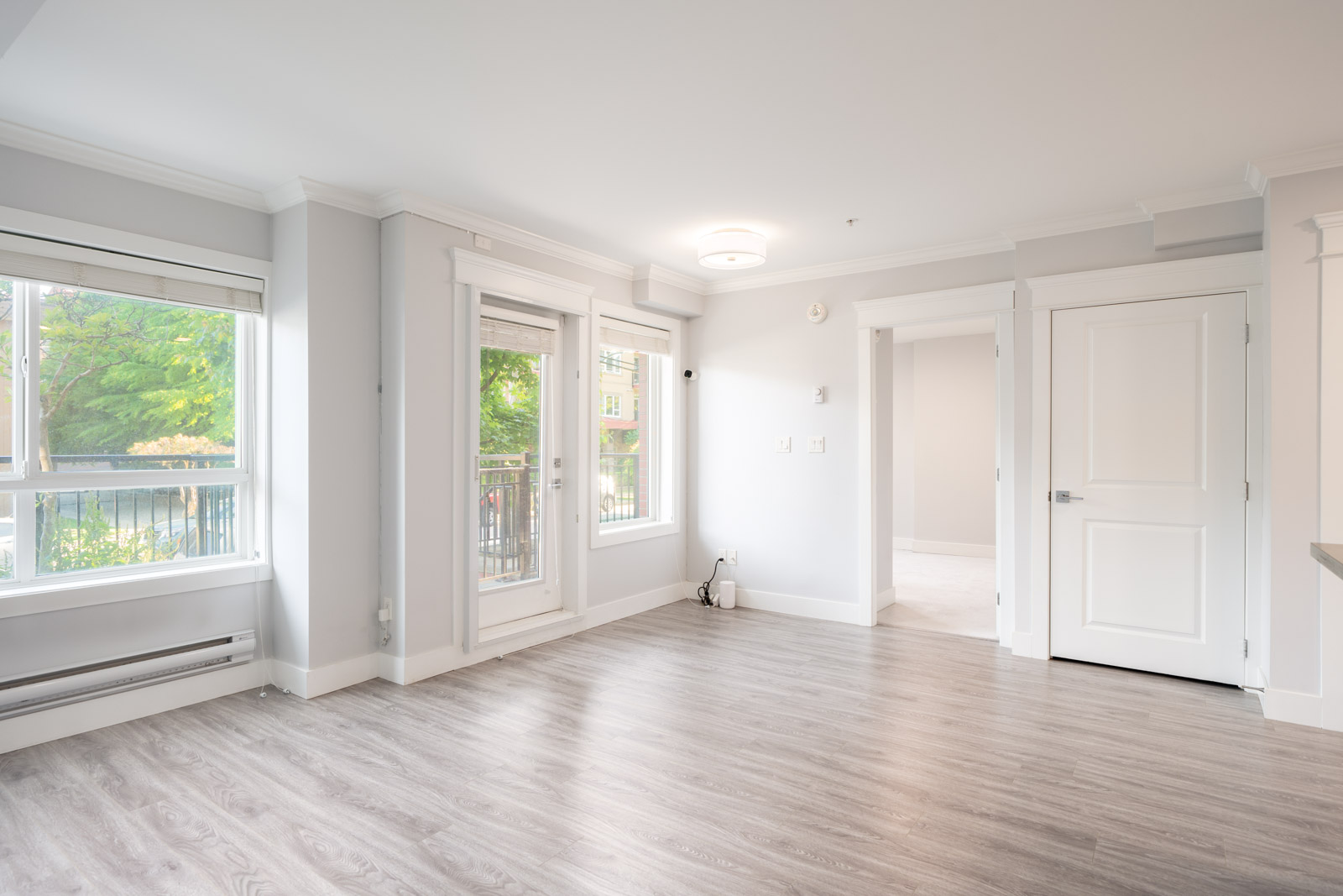 A bright, empty room with large windows, glass doors leading to a balcony, light gray walls, and wood-style flooring.