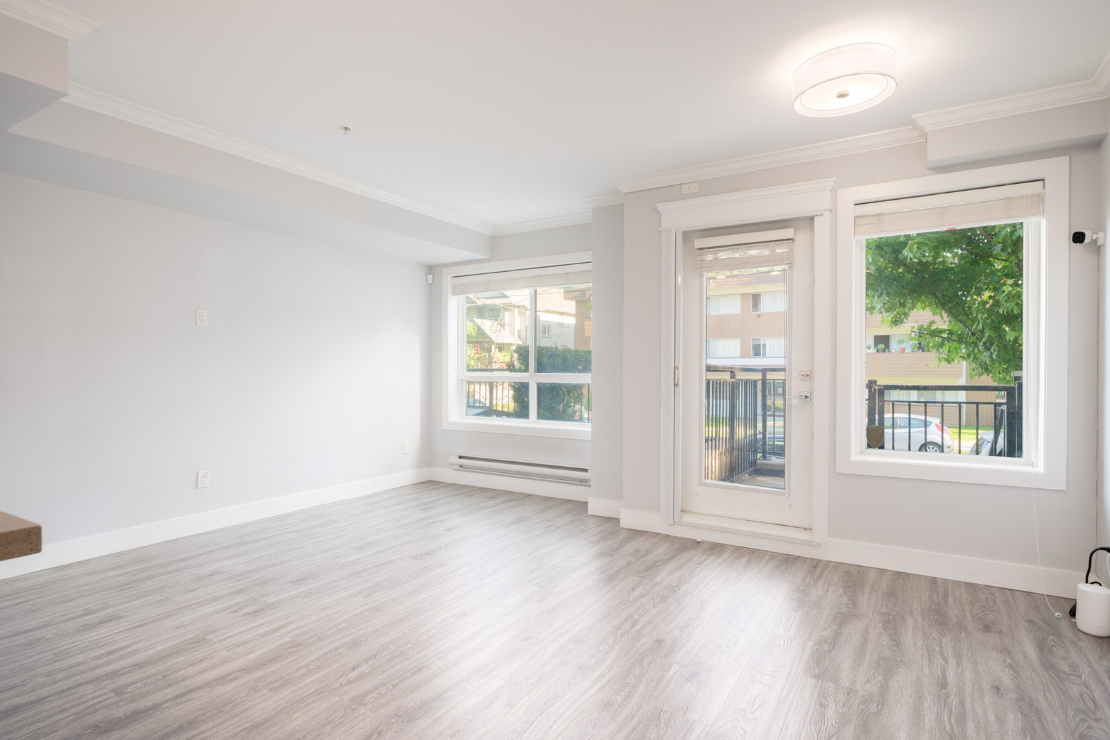 Bright, empty living room with light gray walls, large windows, glass door to balcony, and wood-style flooring.