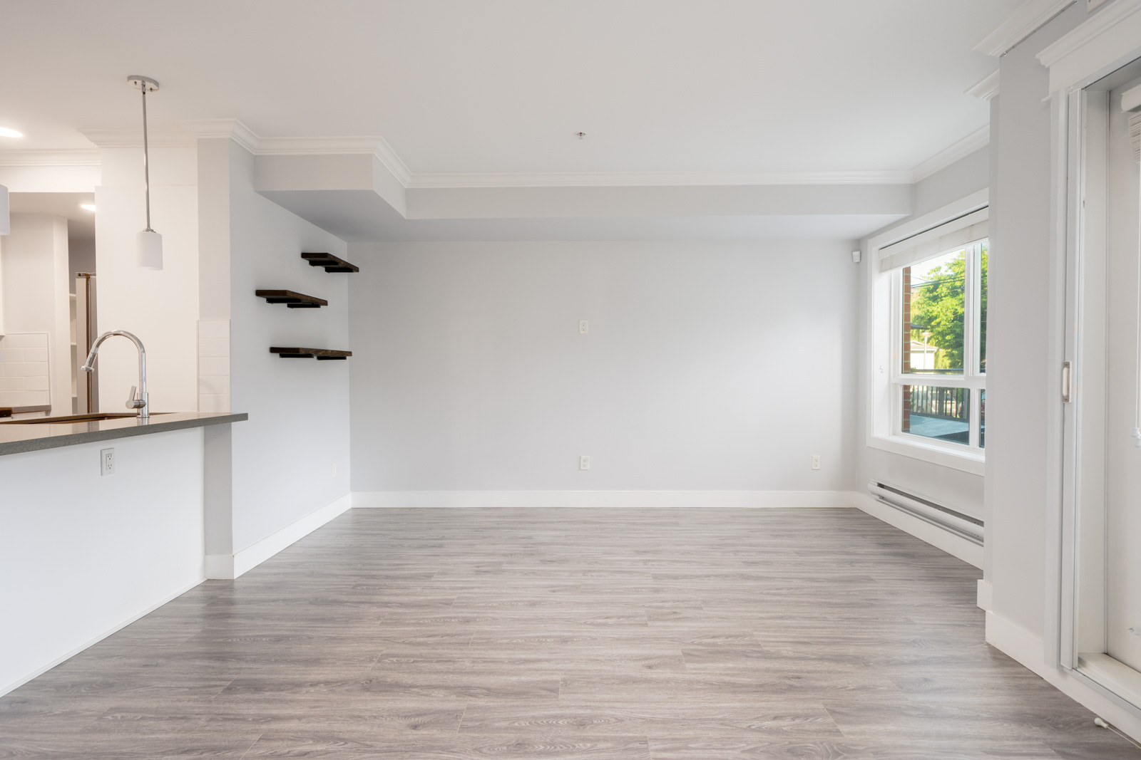 Unfurnished room with light gray walls, wood-look flooring, a kitchen counter with a sink on the left, wall shelves, large window, and sliding glass door.