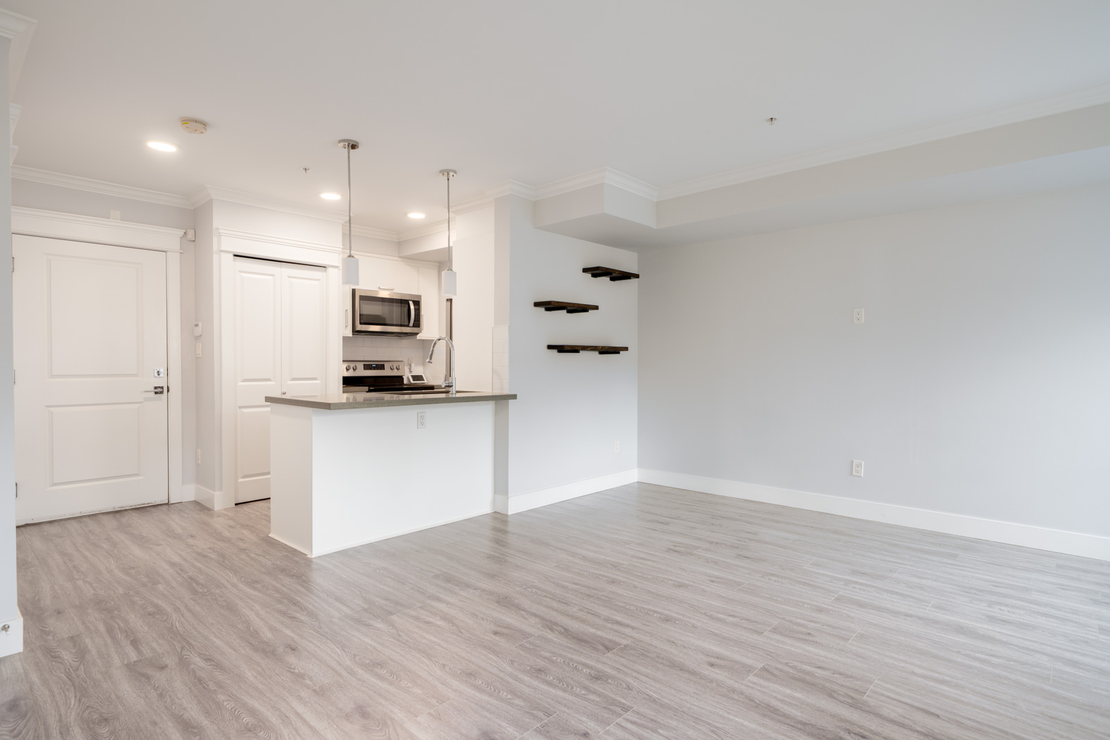 Modern empty apartment with light gray floors, white walls, a small kitchen area with stainless steel appliances, breakfast bar, and floating shelves on one wall.
