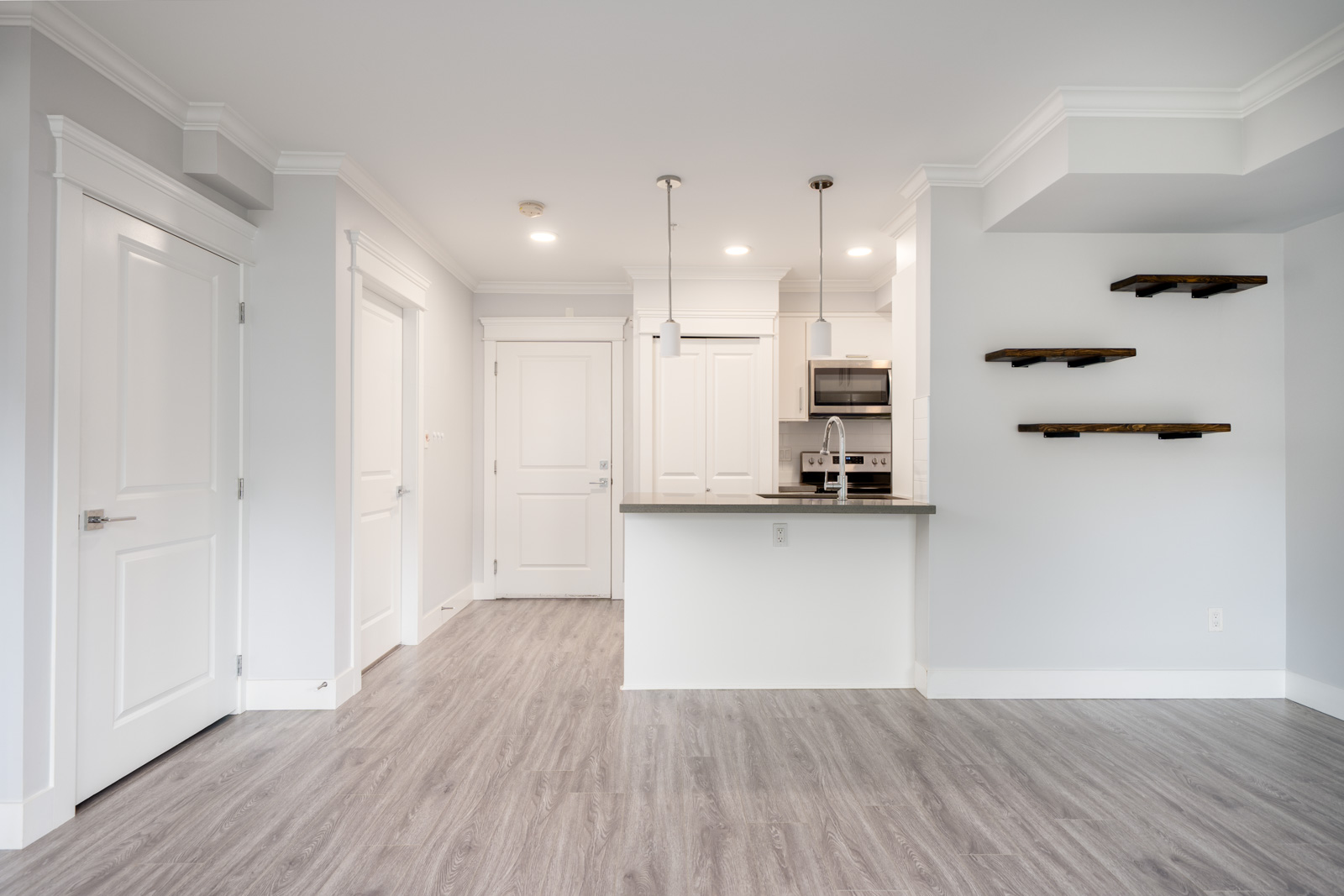 Modern kitchen and entryway with white cabinets, stainless steel appliances, light gray walls, wood-style flooring, and floating shelves on the right wall.