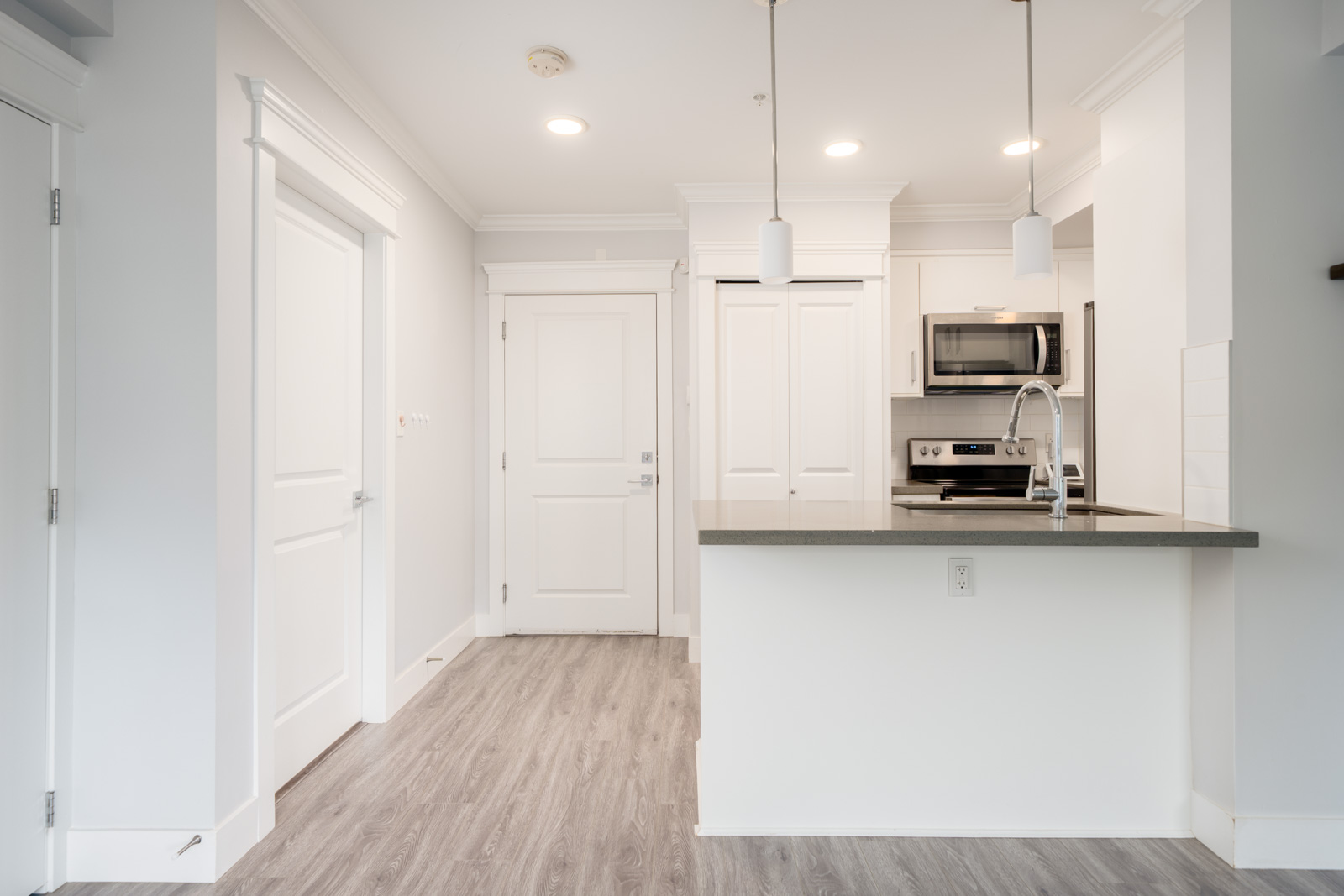 Modern kitchen with white cabinets, stainless steel appliances, pendant lights, a gray countertop, and light wood flooring. A hallway with two doors is visible on the left.
