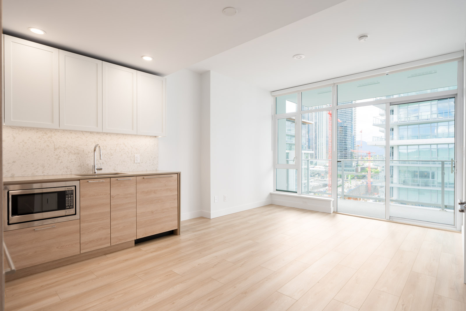 Modern apartment interior with light wood flooring, a small kitchen area with white cabinets and built-in microwave, large windows, and a glass door leading to a balcony with city views.