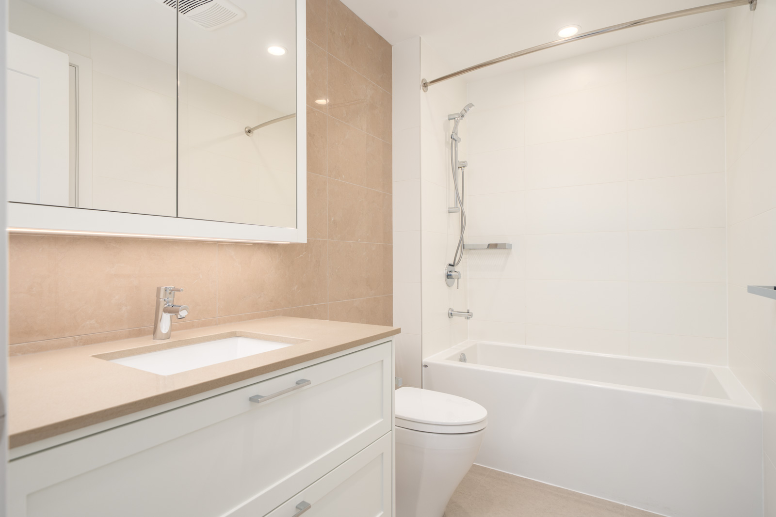 Modern bathroom with a white vanity, sink, toilet, large mirror cabinet, and a bathtub with a showerhead, all in neutral tones and bright lighting.