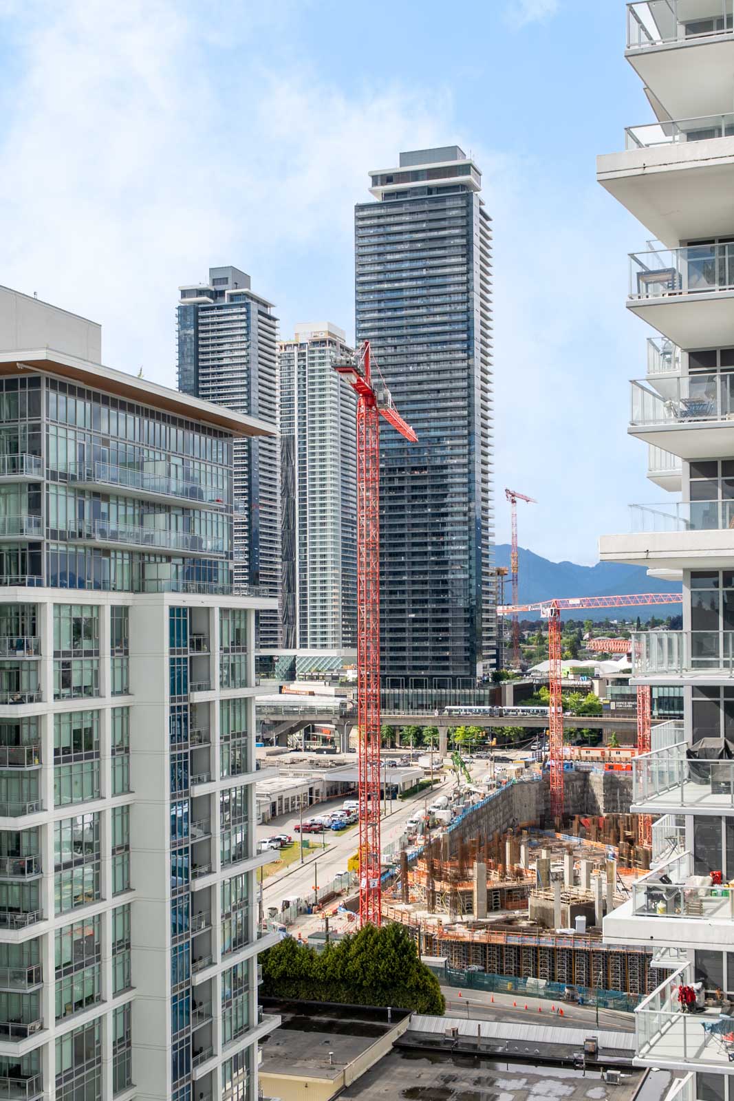 High-rise buildings and construction cranes surround an active construction site in an urban area, with mountains visible in the background.