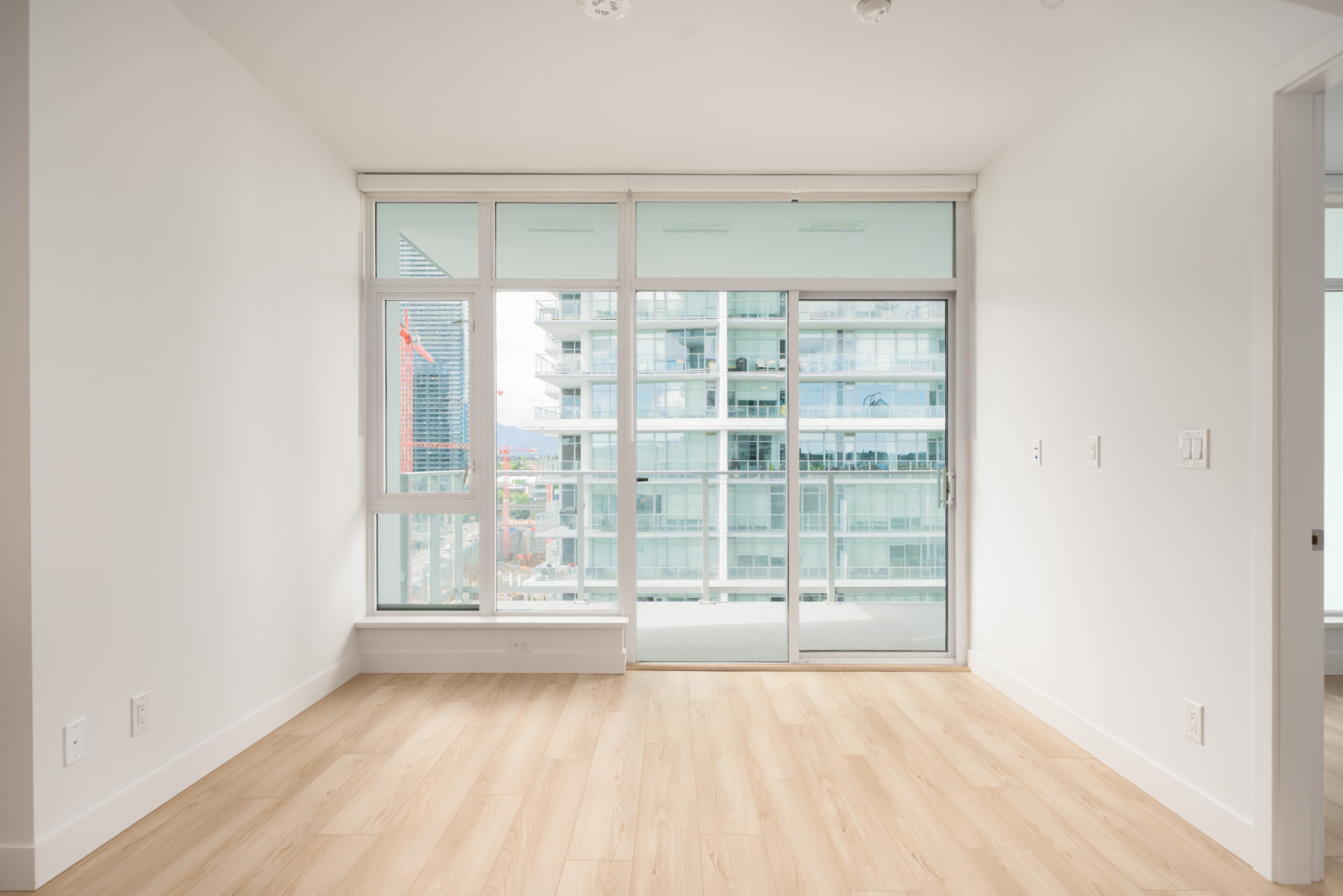 Unfurnished modern apartment room with light wood flooring, white walls, and large floor-to-ceiling windows leading to a balcony overlooking a cityscape.