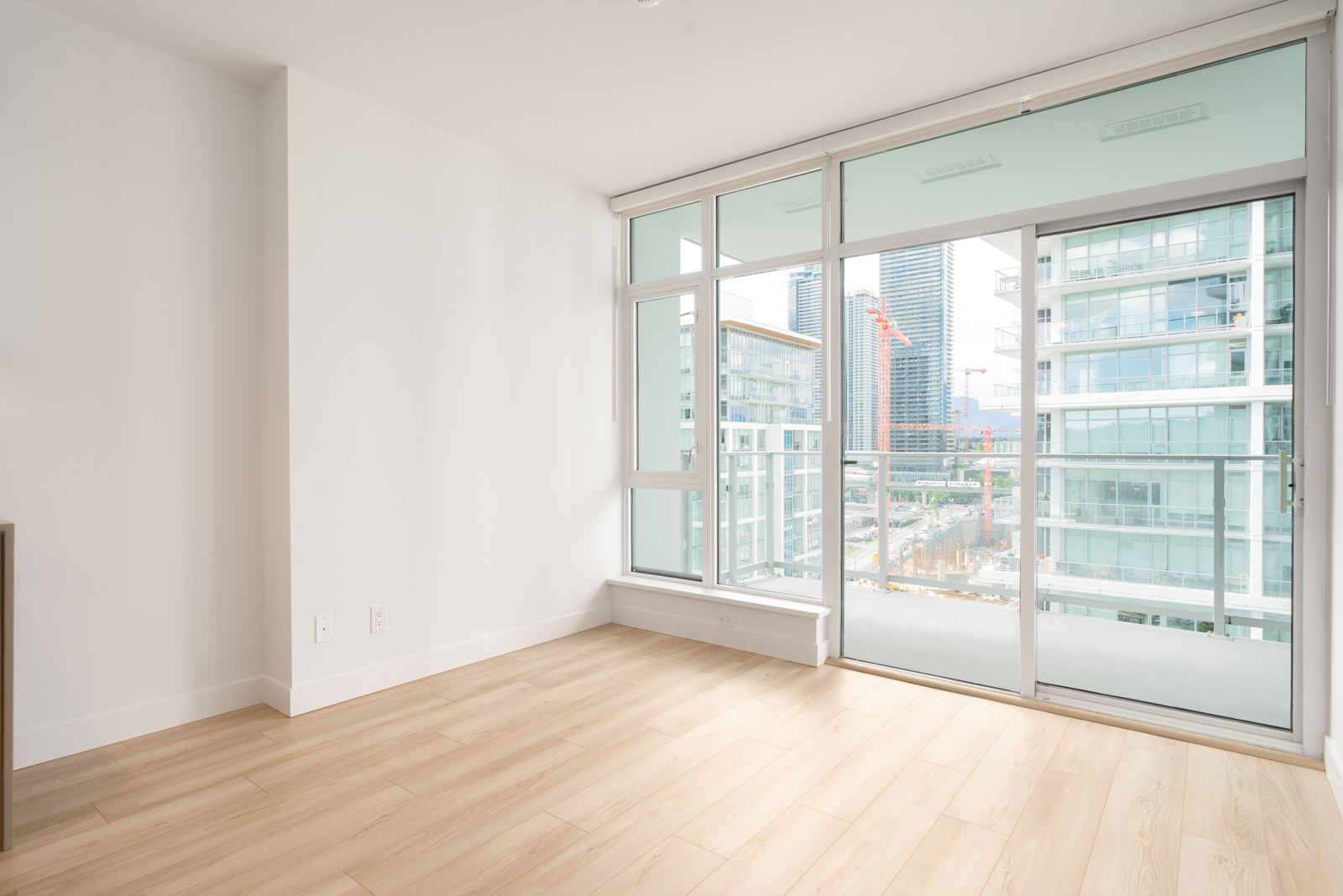 Bright, empty modern apartment room with light wood floors, white walls, and large windows overlooking city buildings and a balcony.