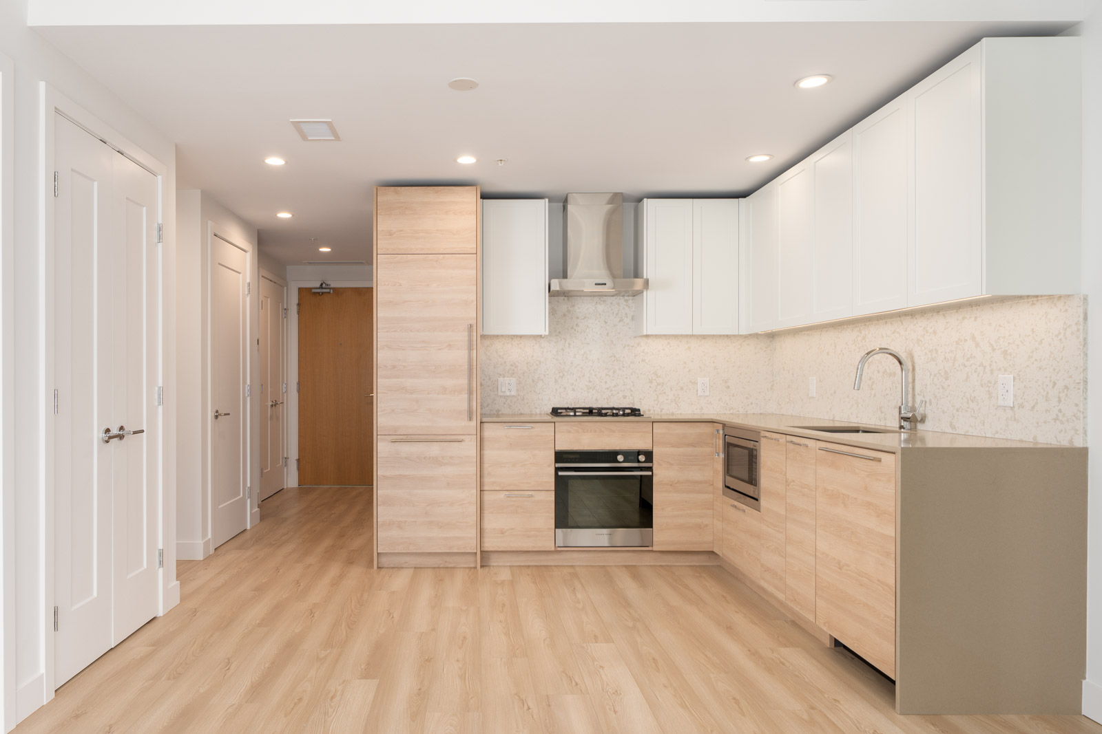 Modern kitchen with light wood cabinets, white upper cabinets, stainless steel appliances, and light wood flooring in an open, unfurnished space.