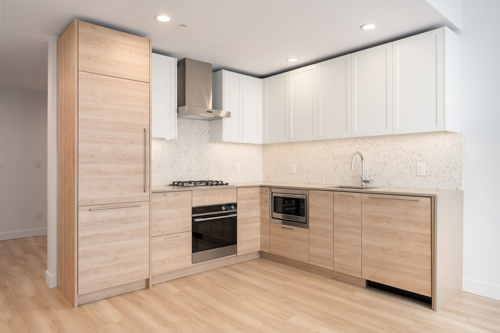 Modern kitchen with light wood cabinets, white upper cabinets, built-in appliances, gas stove, stainless steel range hood, and light-colored backsplash and flooring.
